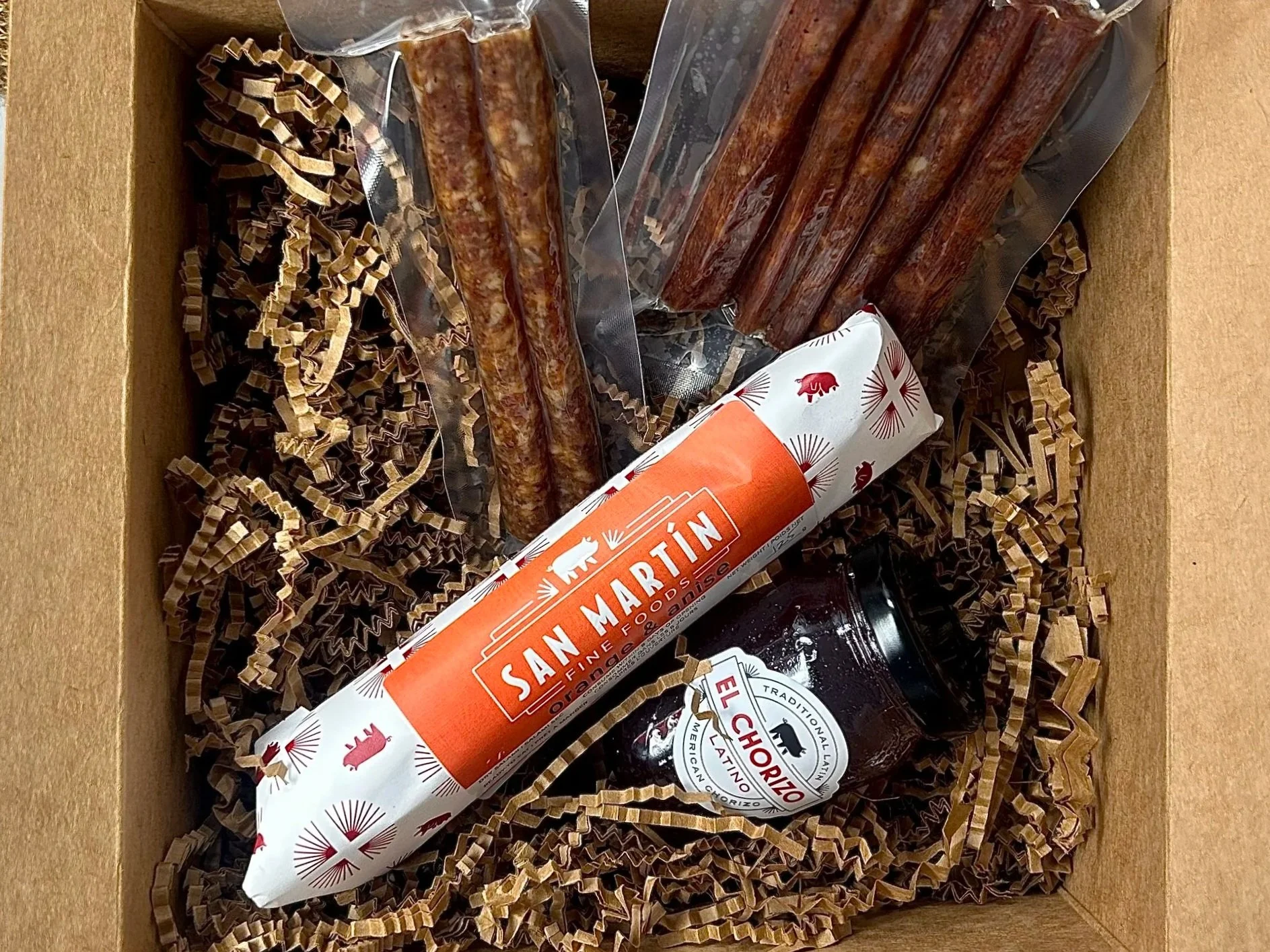 A gift box containing traditional Mexican food items, including a chorizo sausage, a container of mole sauce, and several strips of Mexican chorizo, nestled in brown shredded paper.