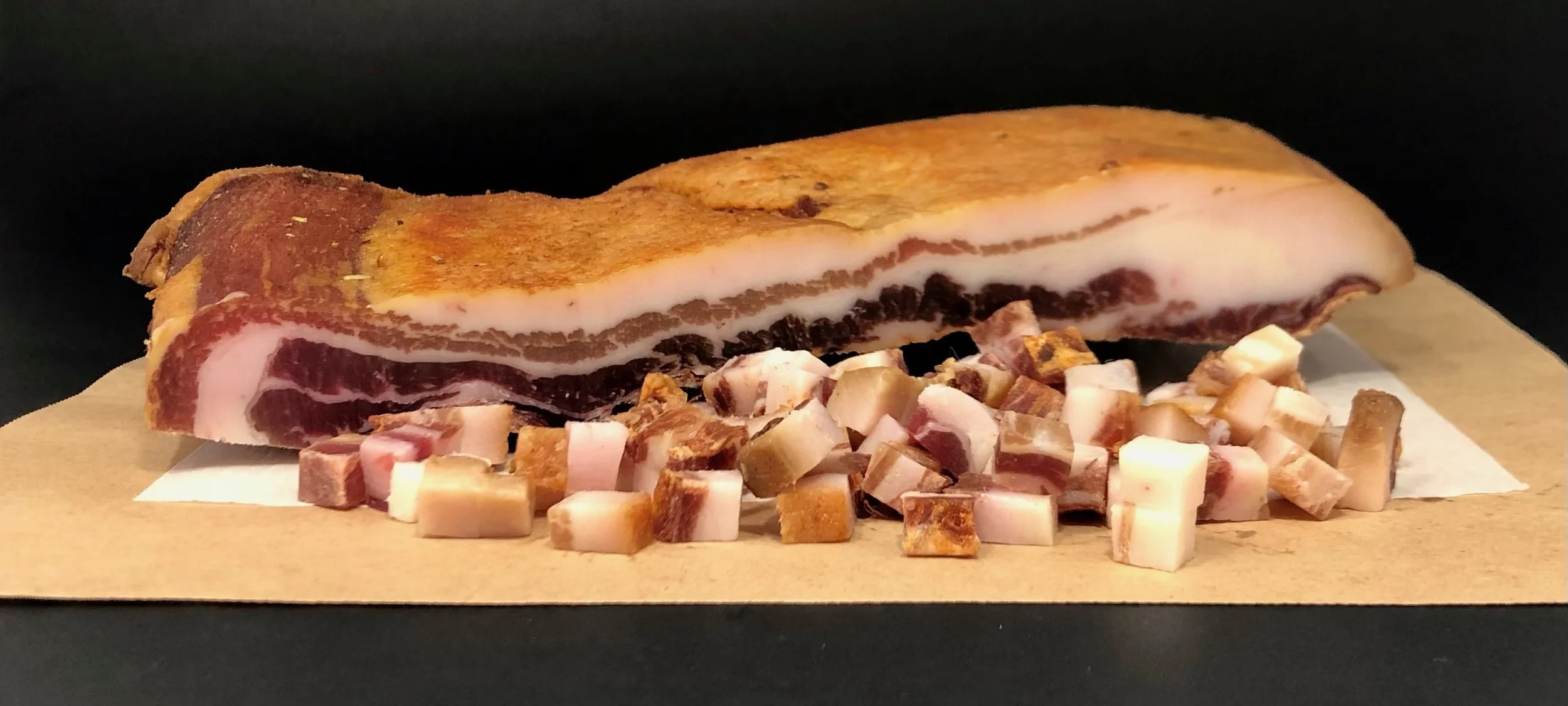 A raw slab of pork belly with layers of meat and fat, placed on a wooden board, surrounded by small diced pieces of pork belly.