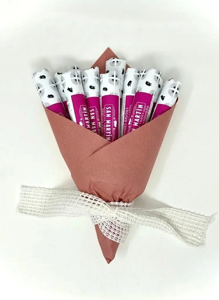 A bouquet made of peppermint candy sticks wrapped in white paper with black fish and abstract designs, arranged in pink wrapping paper tied with a white mesh ribbon. Salami bouquet. salami aesthetic. Valentines day charcuterie. Valentines day.