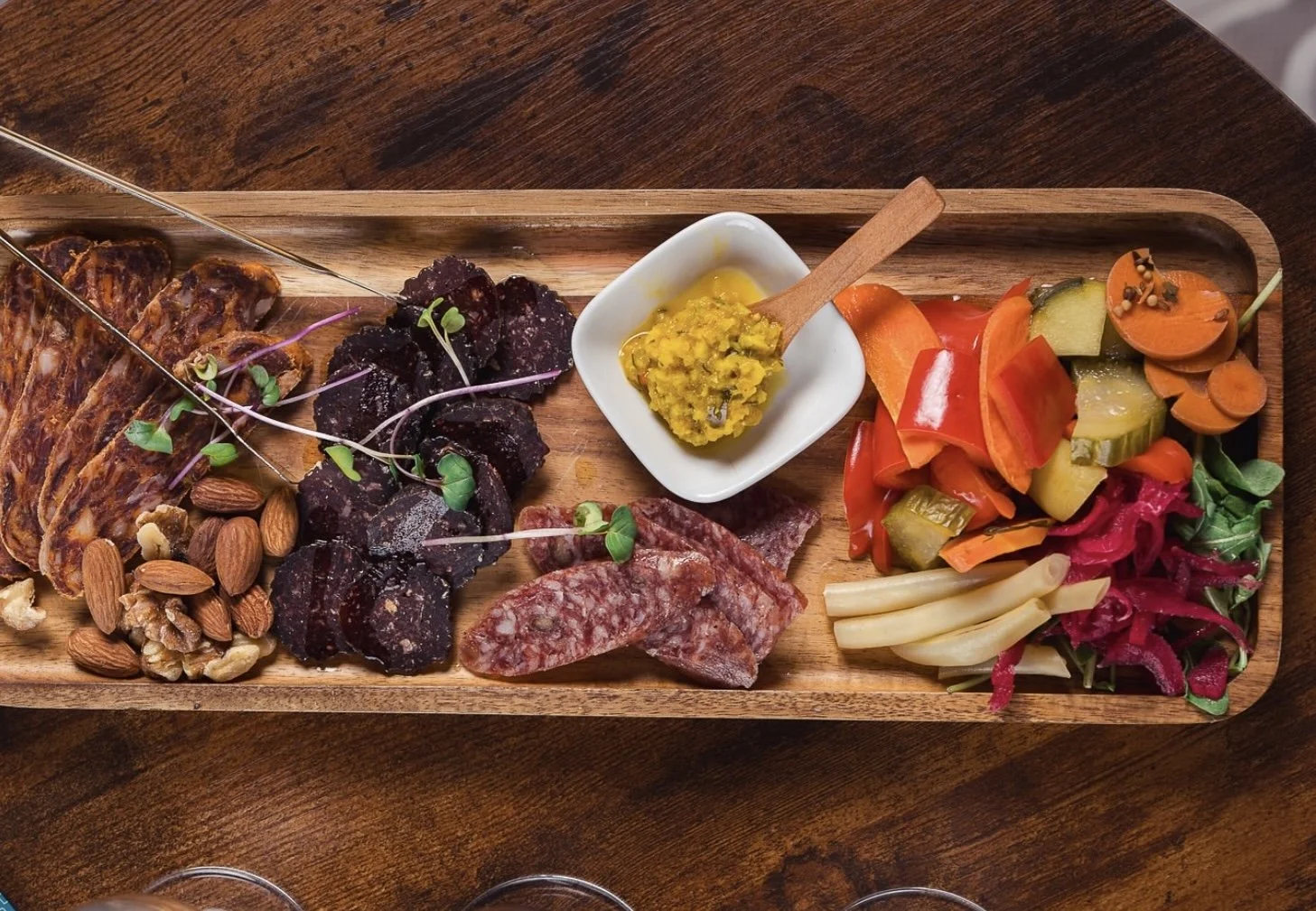 A wooden charcuterie board with slices of cured meats, nuts, and black crackers garnished with microgreens, a small dish of sauce, and a colorful assortment of pickled vegetables including carrots, cucumbers, red peppers, and onions.