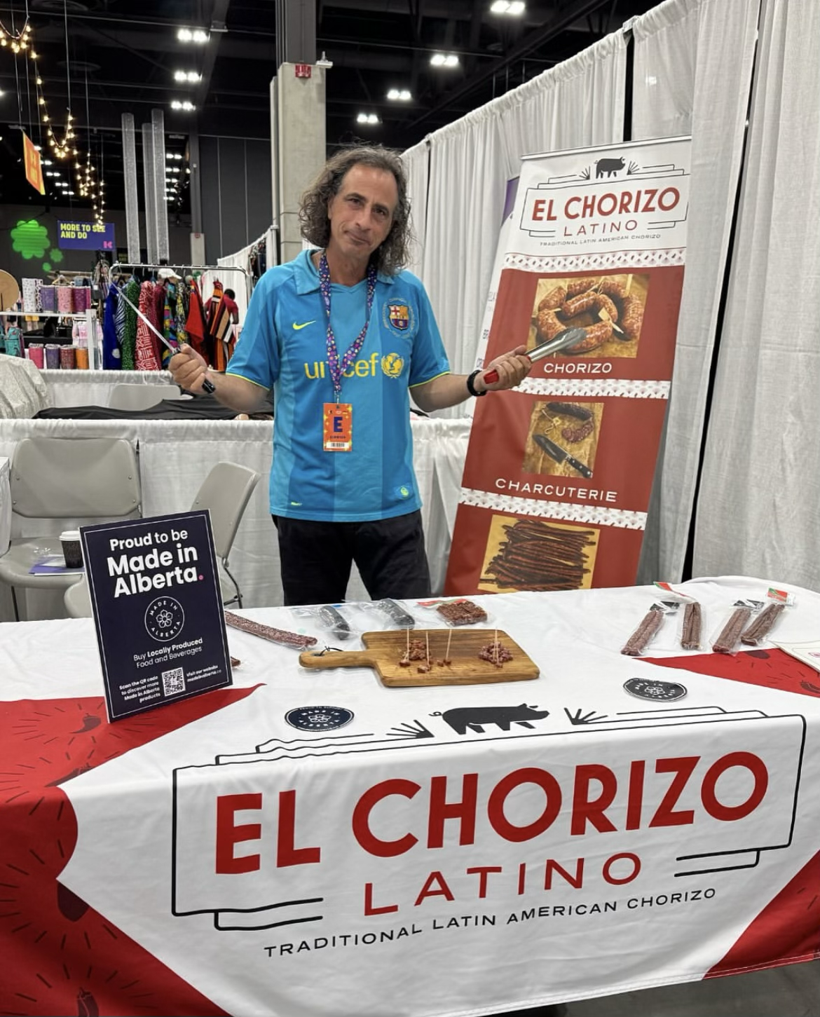 A man standing behind a table at a food booth, holding tongs, with a sign that says "El Chorizo Latino". The table displays different types of chorizo and other meats, with a smaller sign promoting local Alberta products. The booth has a large banner, and the man is wearing a blue Barcelona football jersey and blogging or event accreditation tags.