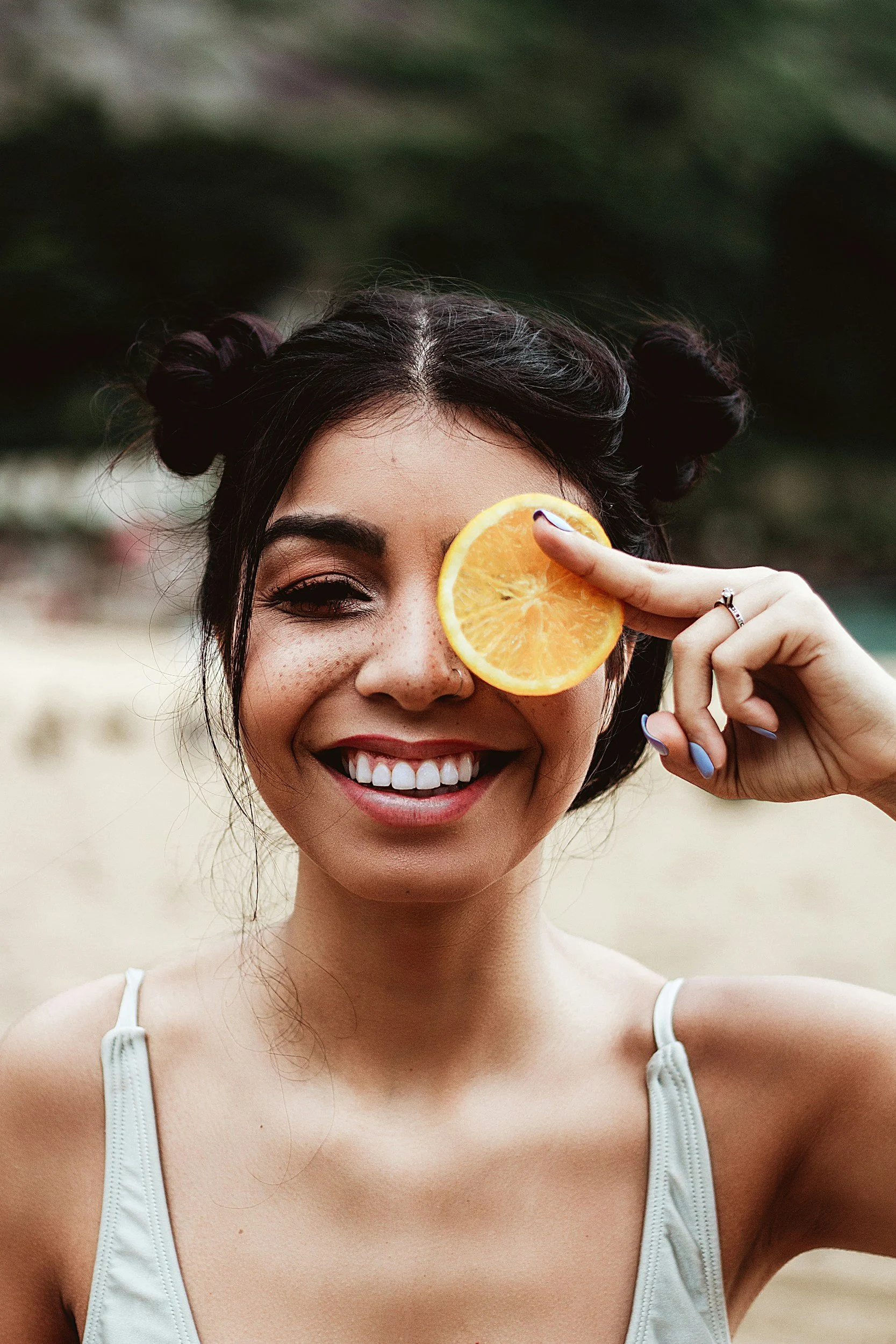 A woman with dark hair styled in buns, smiling, holding a slice of orange over her right eye, outdoors, beach background.