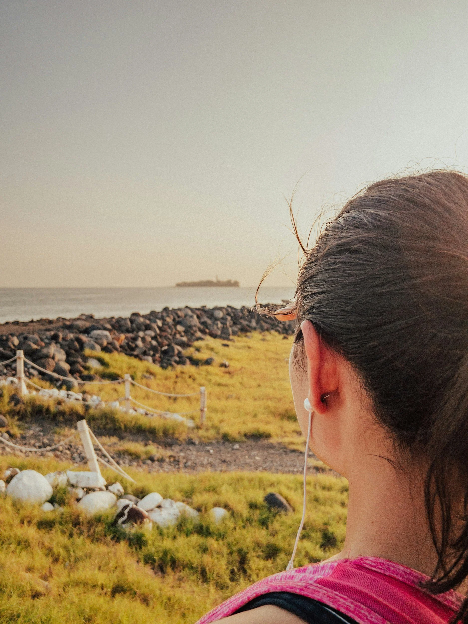 A woman with dark hair in a ponytail wearing a pink shirt and white earphones, looking towards a rocky shoreline and calm ocean during sunset.