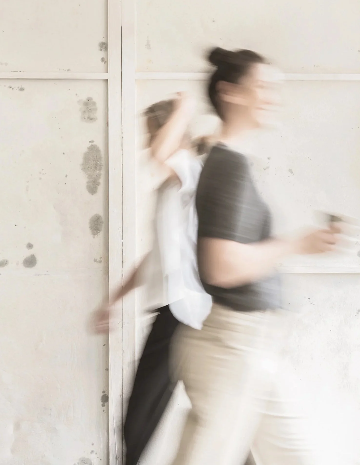 Grafikerin Melanie Pototschnig laeuft lachend von links nach rechts durch das Bild mit einer Tasse Kaffee in der Hand. In die Gegenrichtung laeuft Texterin Gertrud Kainz. Das Bild ist unscharf und in einem warmen beige Ton.