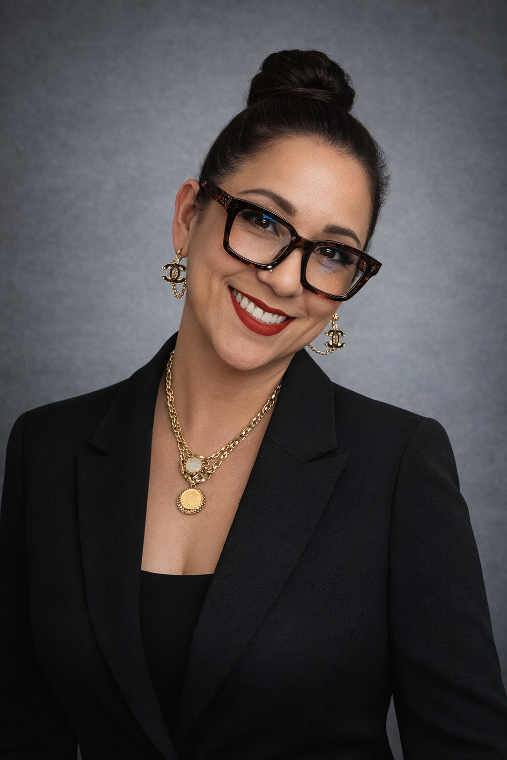 A professional woman with dark hair in a bun, wearing large tortoiseshell glasses, gold jewelry, red lipstick, and a black blazer, smiling at the camera.