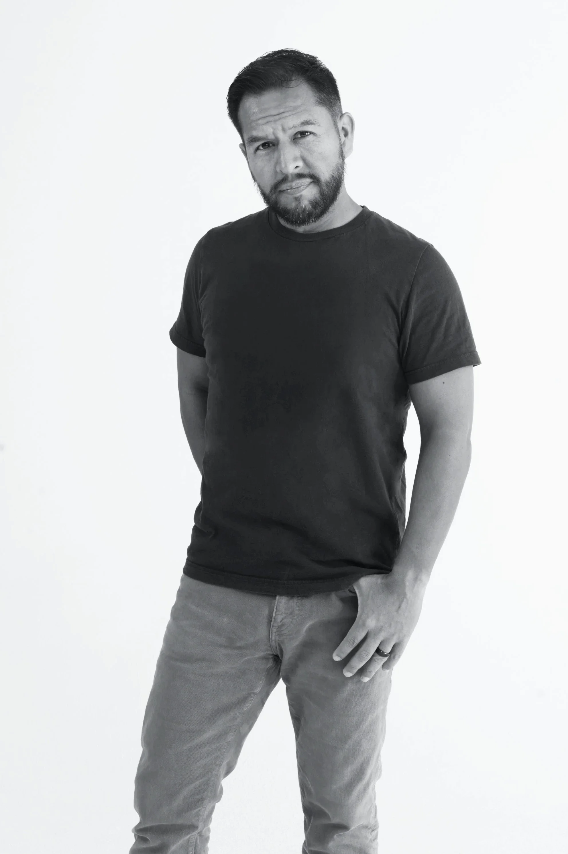 A man with a beard and short dark hair wearing a black t-shirt and gray pants, standing against a plain light background, looking at the camera with a slight tilt of his head and a serious expression.