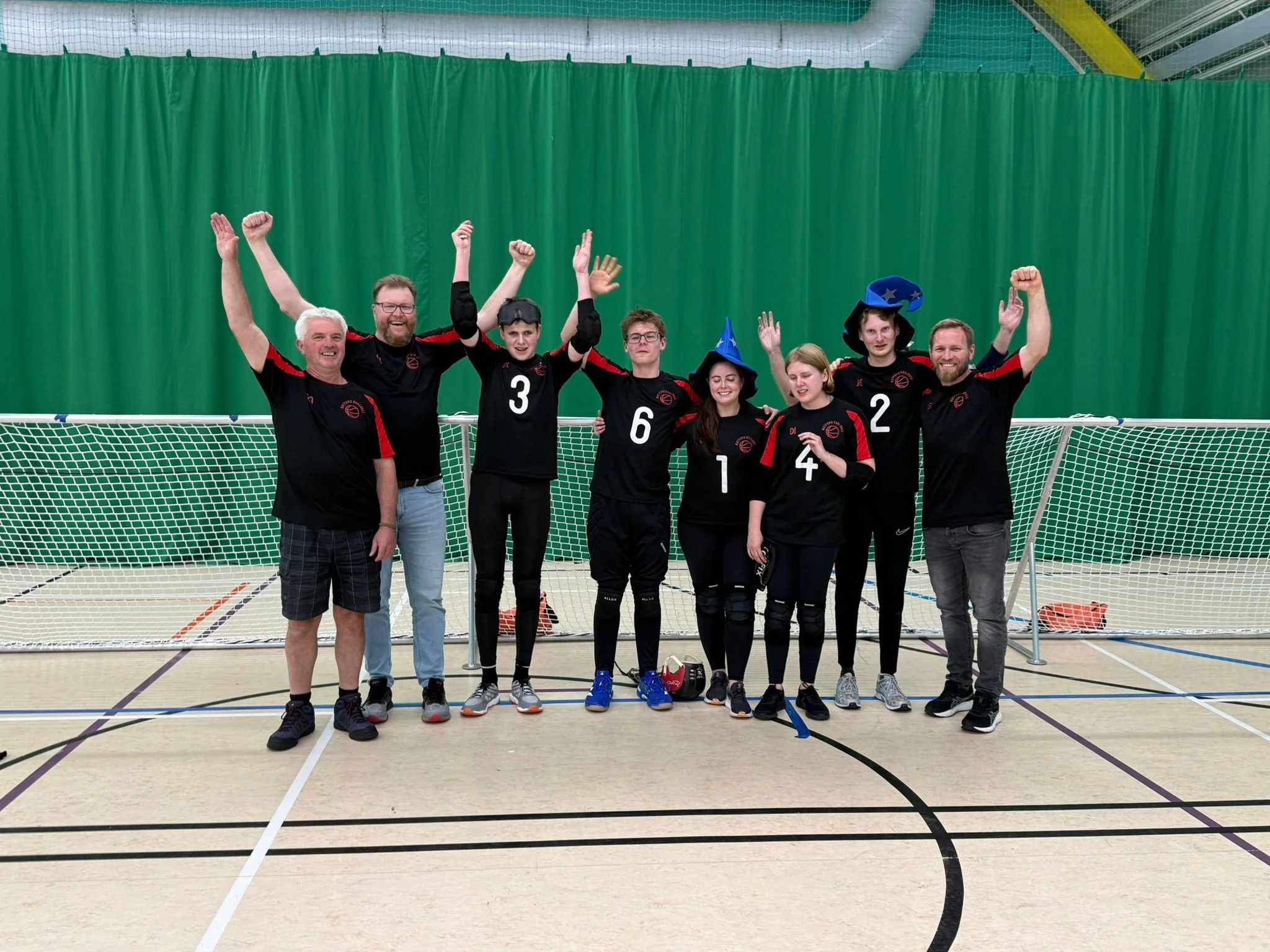   ALT text:  Watford Wizards goalball players and coaches standing together on an indoor sports court, celebrating with arms raised in front of the goalball net. Several players are wearing black Watford Goalball kit with red detailing, and two playe