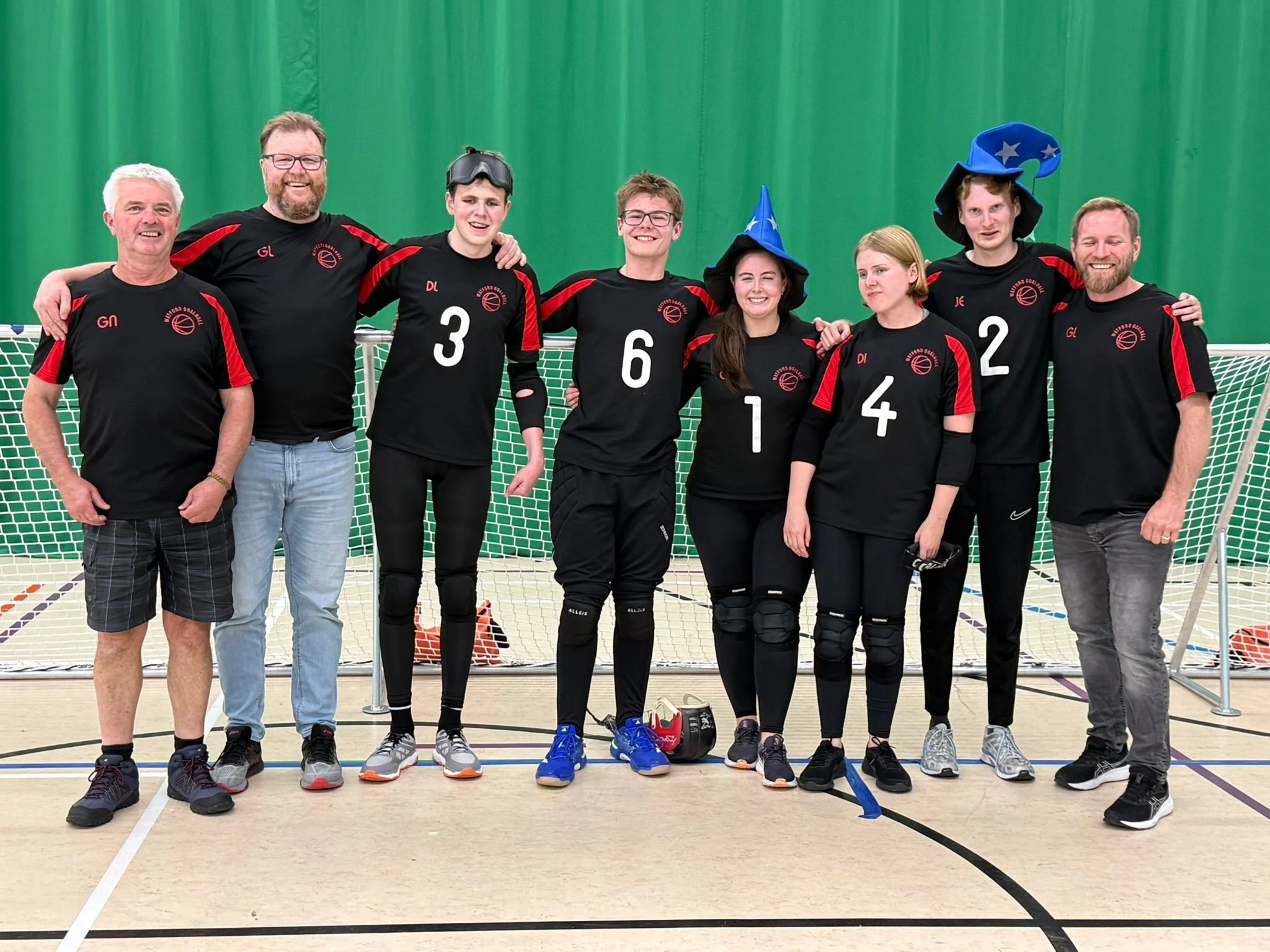   ALT text:  Watford Wizards goalball team photo on an indoor court. Players and coaches stand shoulder to shoulder in front of the goalball net, wearing black and red Watford Goalball kit, with two players wearing blue wizard hats. 