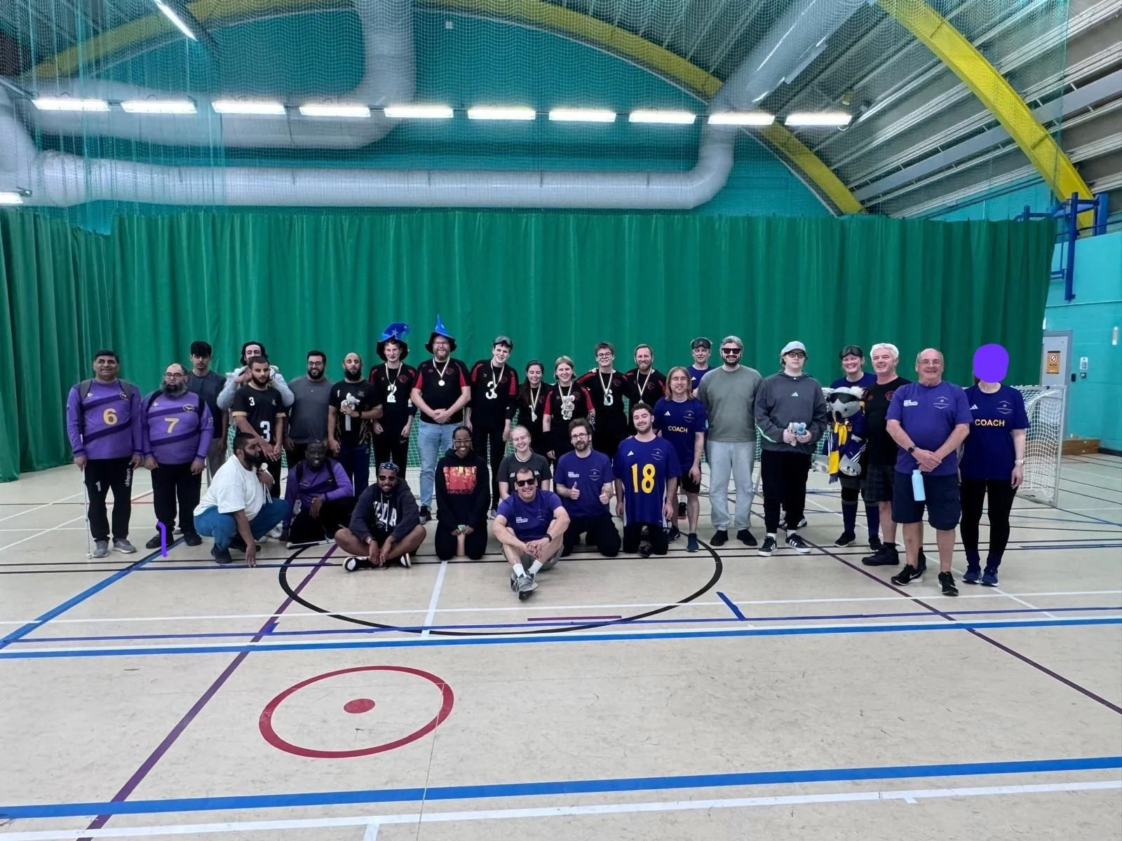   ALT text:  Large group photo of goalball teams, coaches and supporters on an indoor sports court after a tournament. Watford Wizards players are pictured among the group wearing black and red kit, with other teams in purple and dark kits. 