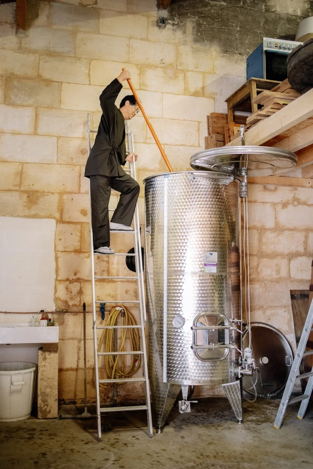 Une personne en vêtements de maison utilisant une longue baguette pour atteindre le sommet d'un grand récipient en acier inoxydable dans un garage ou une cave, avec des murs en pierre et des étagères en bois au-dessus.