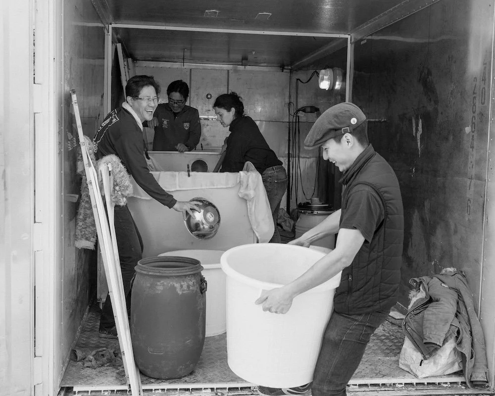 Groupe de personnes en train de laver des objets dans une petite pièce, avec quelques bacs et bidons, ambiance conviviale, en noir et blanc.
