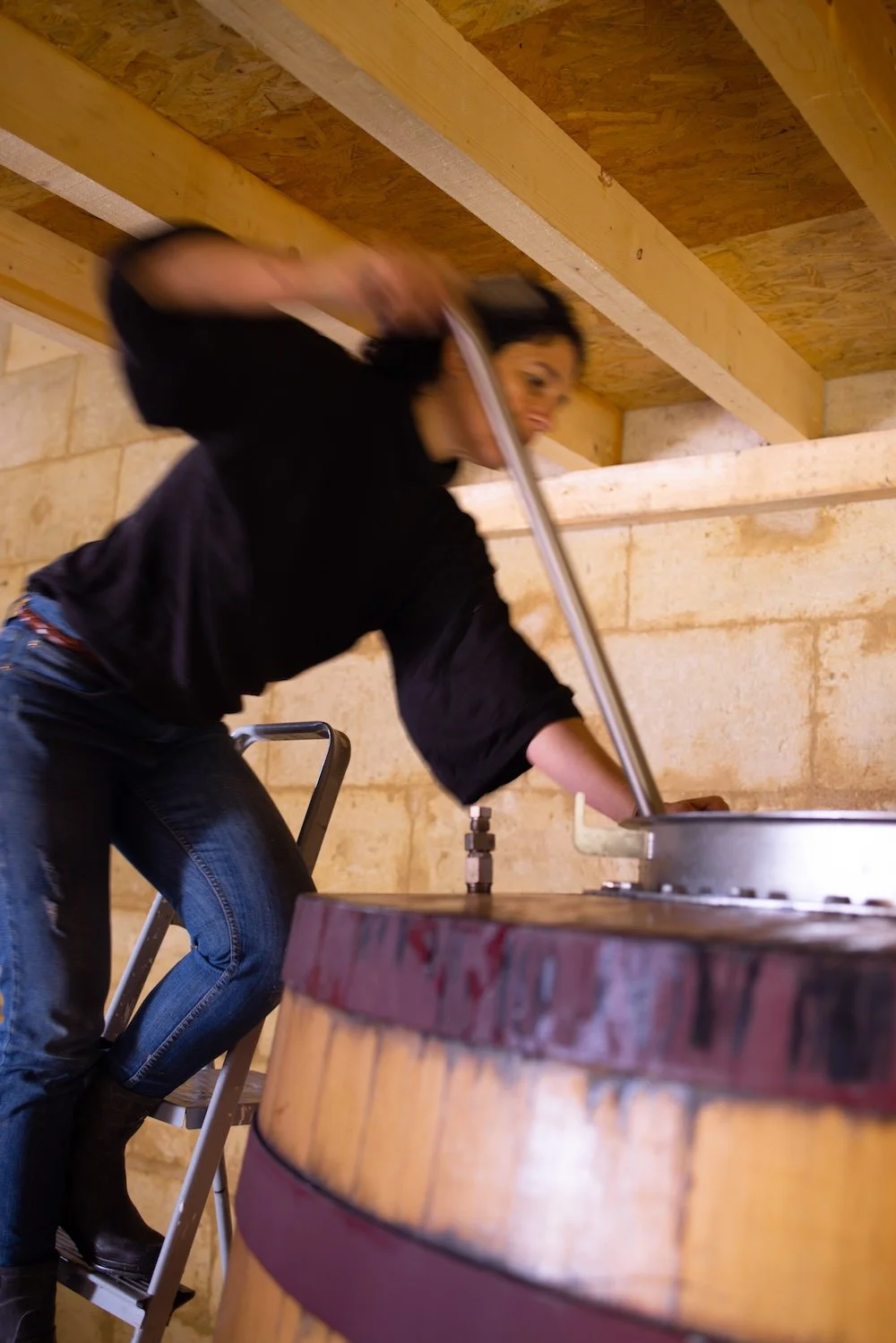 Une femme en train de remuer une cuve en bois dans une cave ou un espace de fermentation, avec des murs en pierre.