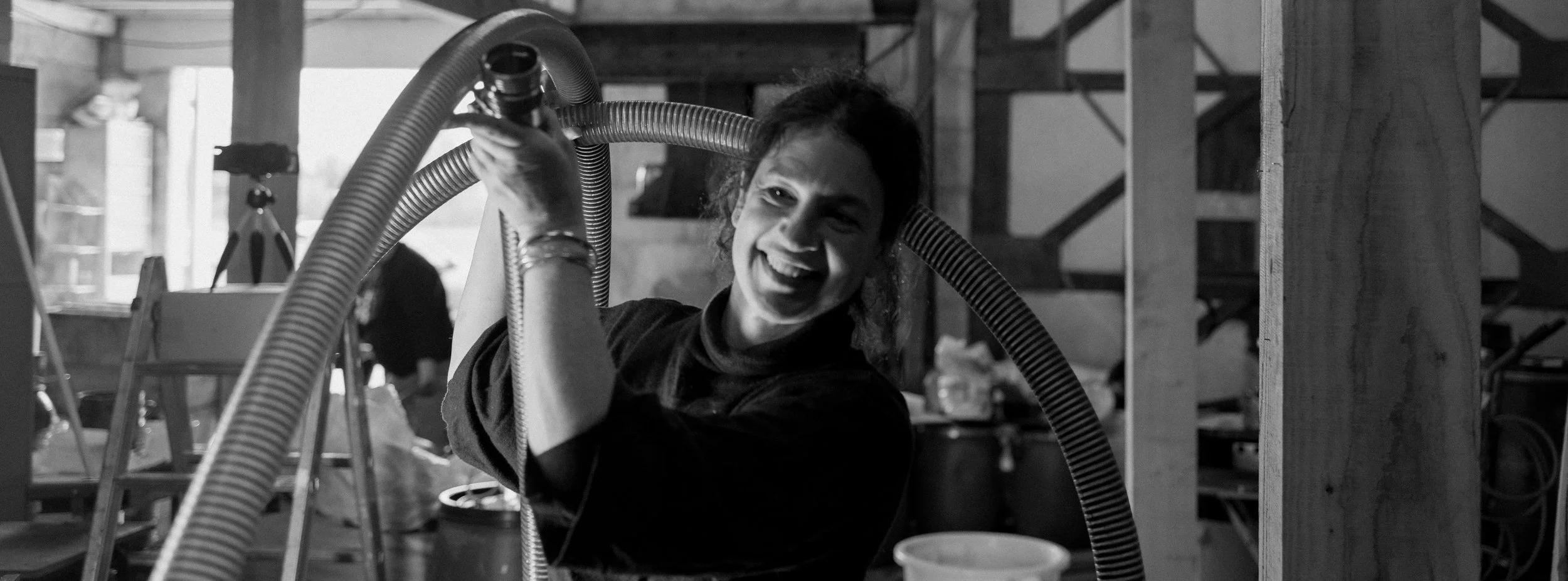 Une femme souriante dans un atelier, tenant un tuyau flexible.