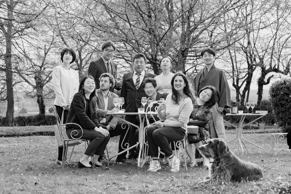 Groupe de neuf personnes posant pour une photo en extérieur avec un chien, certaines tenant des verres de vin, dans un jardin avec des arbres en arrière-plan.