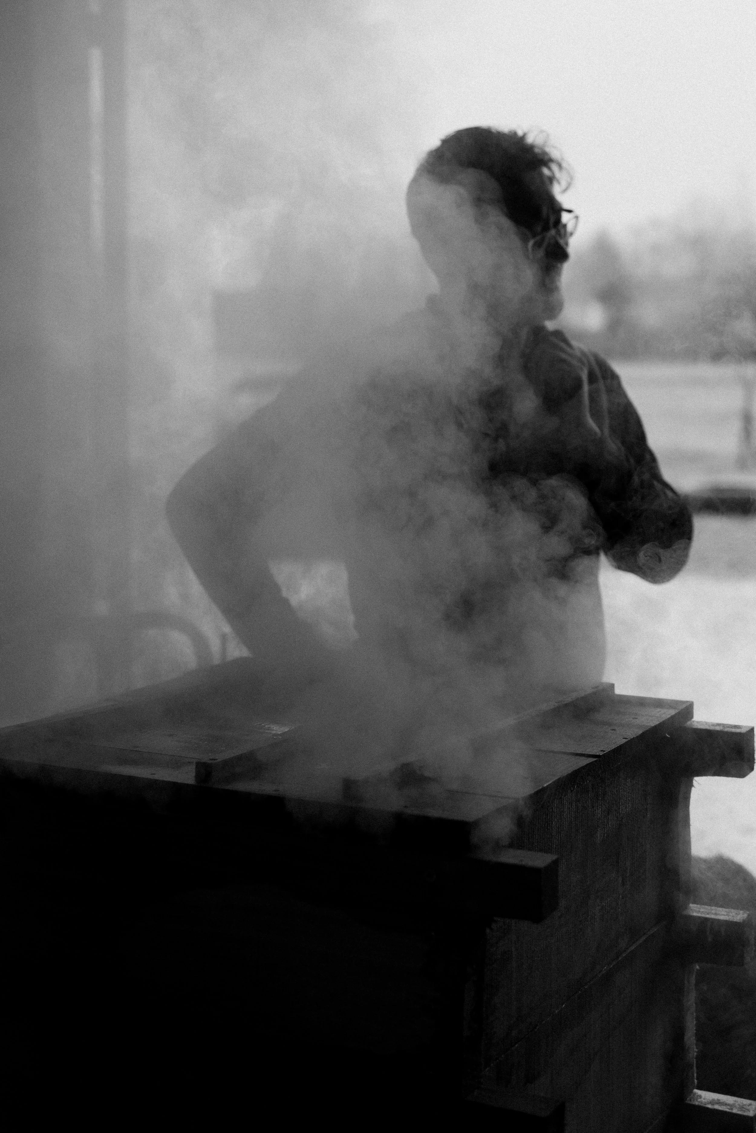 Une personne portant des lunettes et un sweat à capuche, vue en profil, derrière de la vapeur ou de la fumée noire, près d'une table en bois.