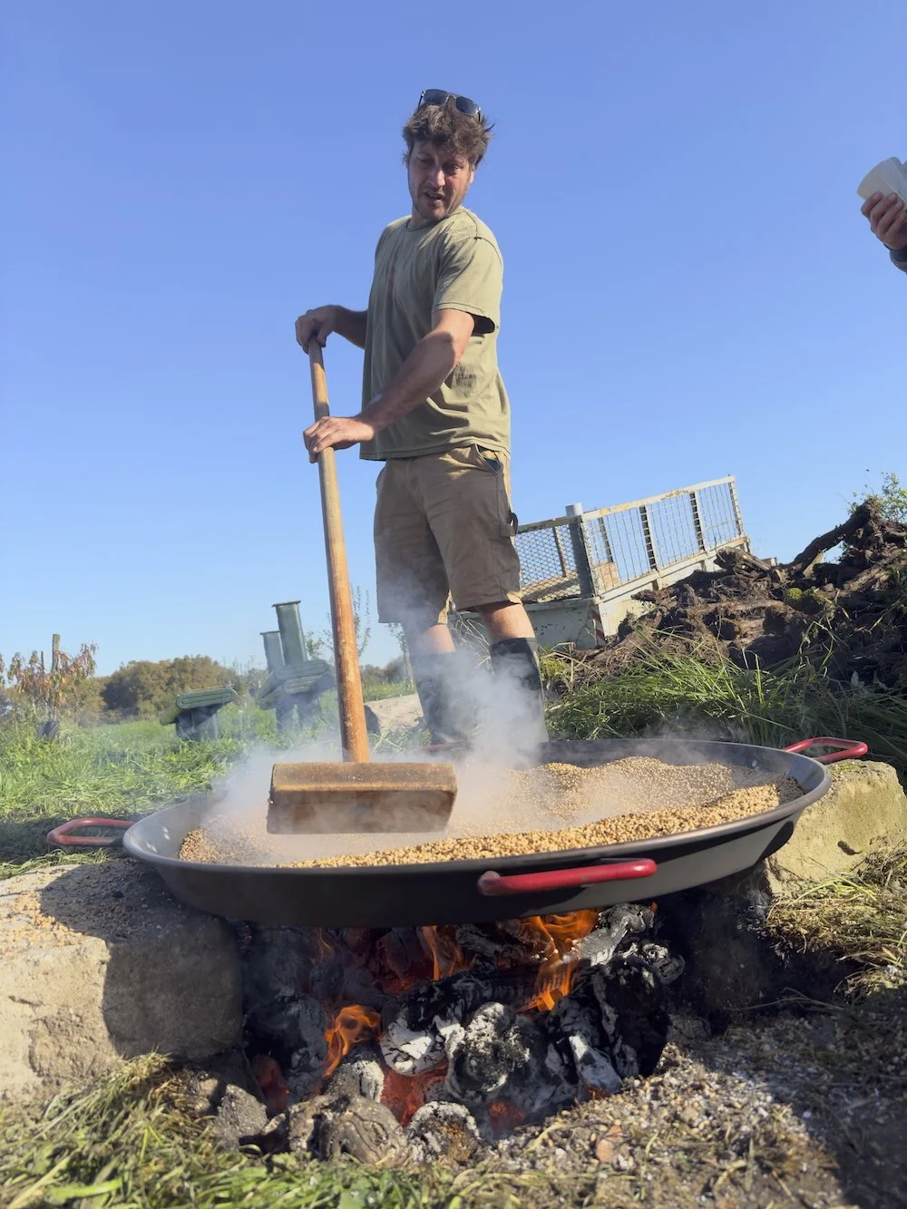 Un homme fait du camping ou de la randonnée, utilisant une pelle pour remuer des lentilles ou du riz dans une grande poêle posée sur un feu de bois en plein air, dans un champ en plein jour.