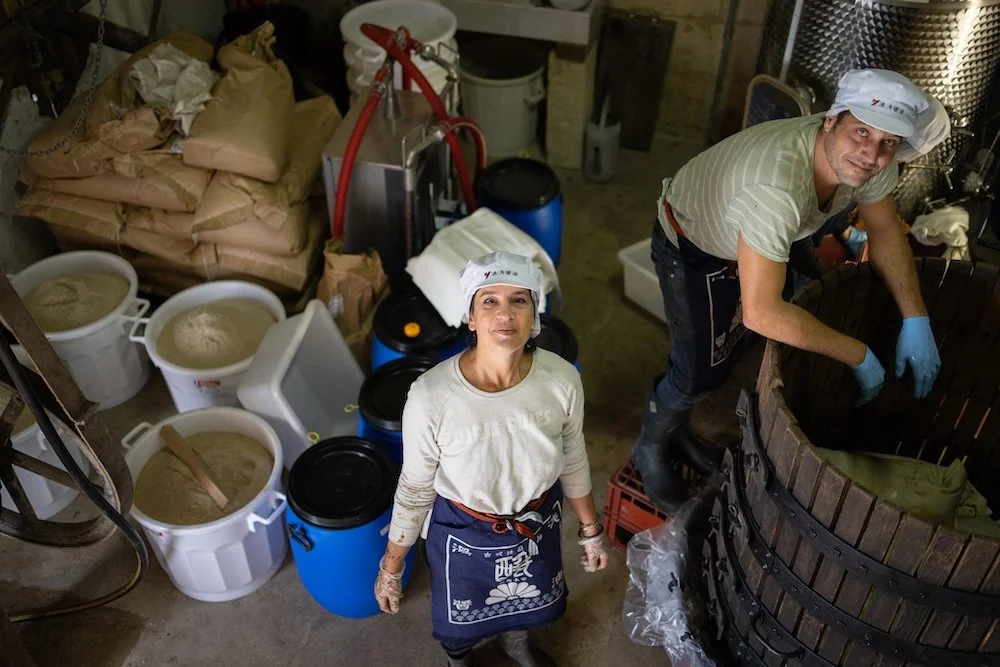 Deux personnes travaillant dans une cave à vin, utilisant des gants en latex, entourées de fûts de vieillissement et de grandes quantités de levure ou de mouture pour la fabrication du vin.