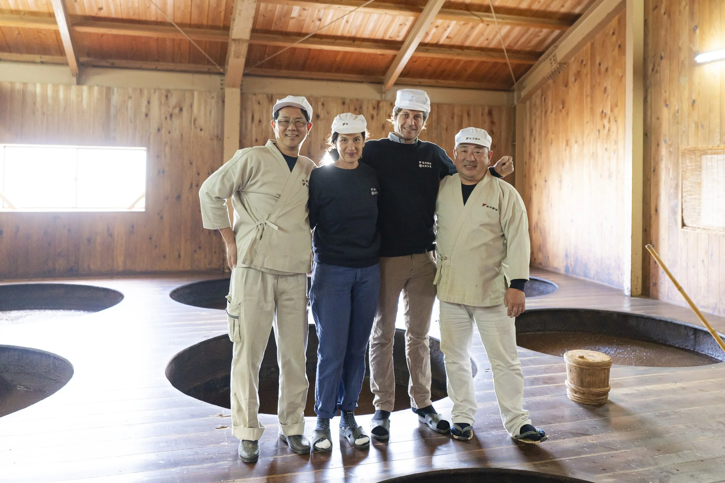 Cinq personnes posant pour une photo dans une salle en bois, certains portant des tenues de chef ou de travail, d'autres en vêtements décontractés, avec des plans d'eau ou de cuisson dans le sol en bois derrière eux.