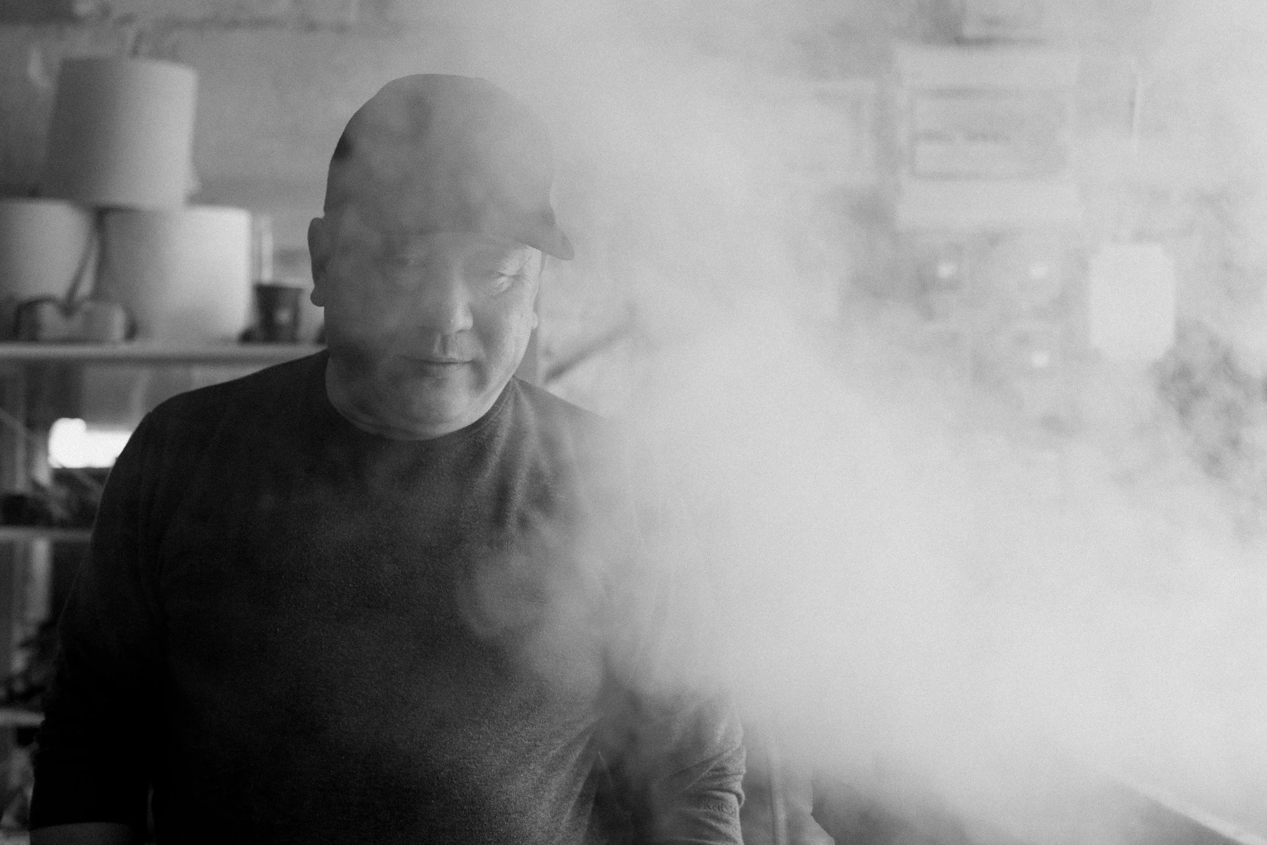 Un homme porte une casquette noire et un pull noir, se trouvant dans une cuisine, avec de la vapeur qui s'élève près de lui.