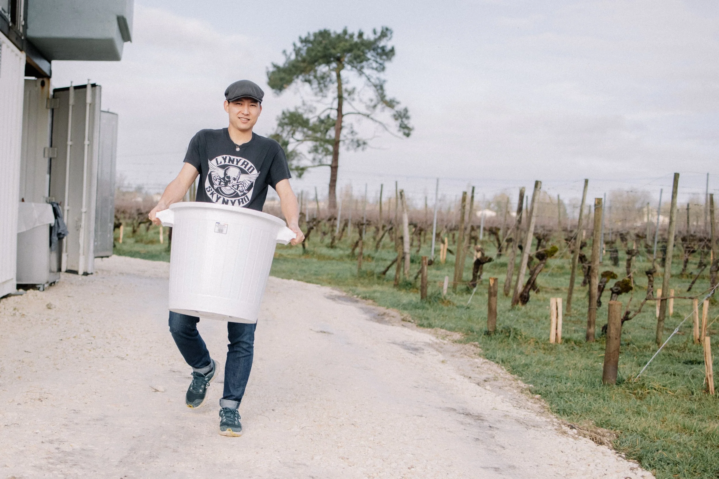 Un homme portant un T-shirt noir avec un dessin de skull et le texte 'Lynyrd Skynyrd', tenant un grand seau blanc, dans un vignoble avec des rangées de vignes et un arbre en arrière-plan.