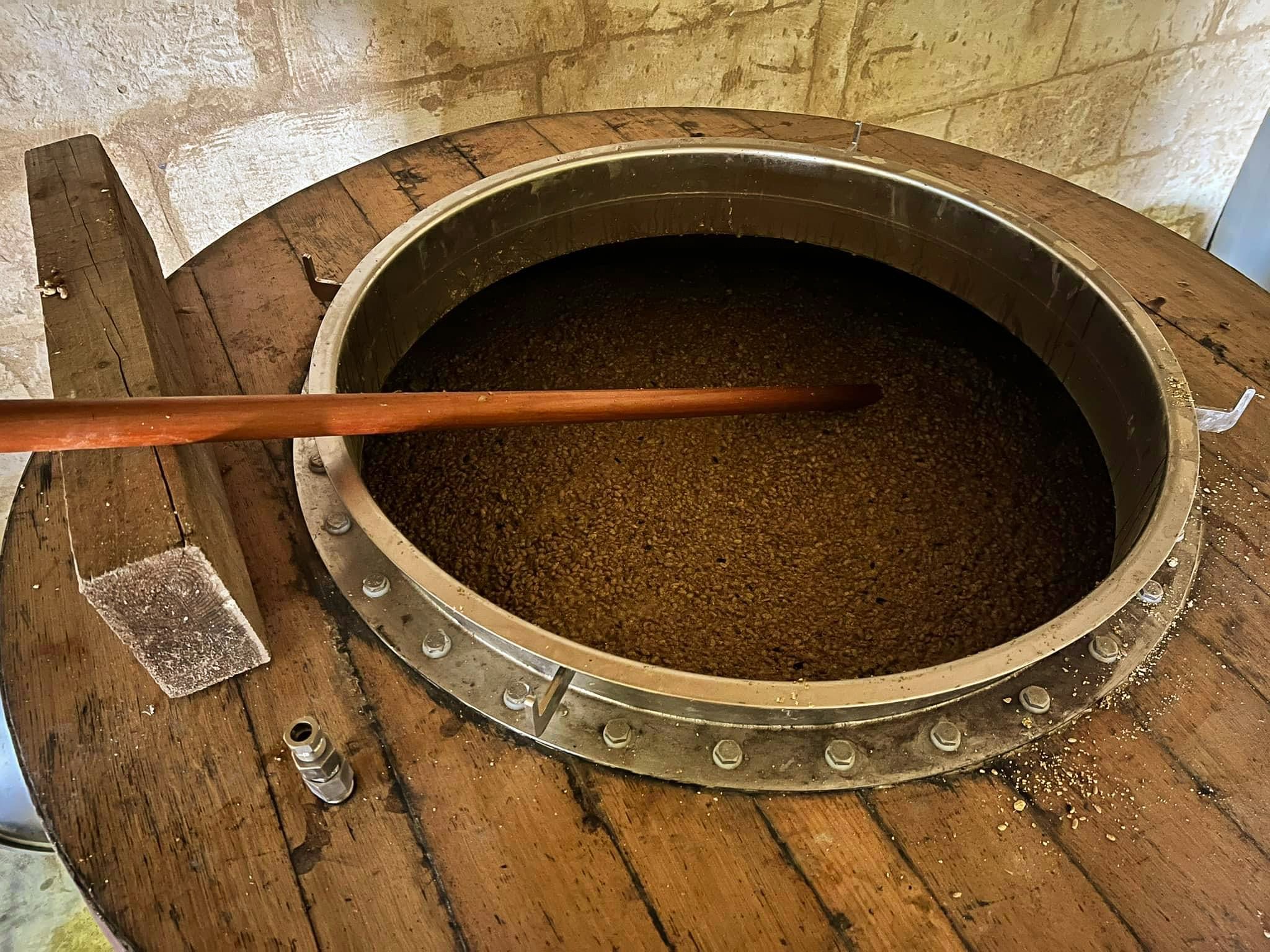 Un grand récipient en métal contenant une boisson sombre, probablement du vin ou du café, avec un long bâton en bois posé à l'intérieur.