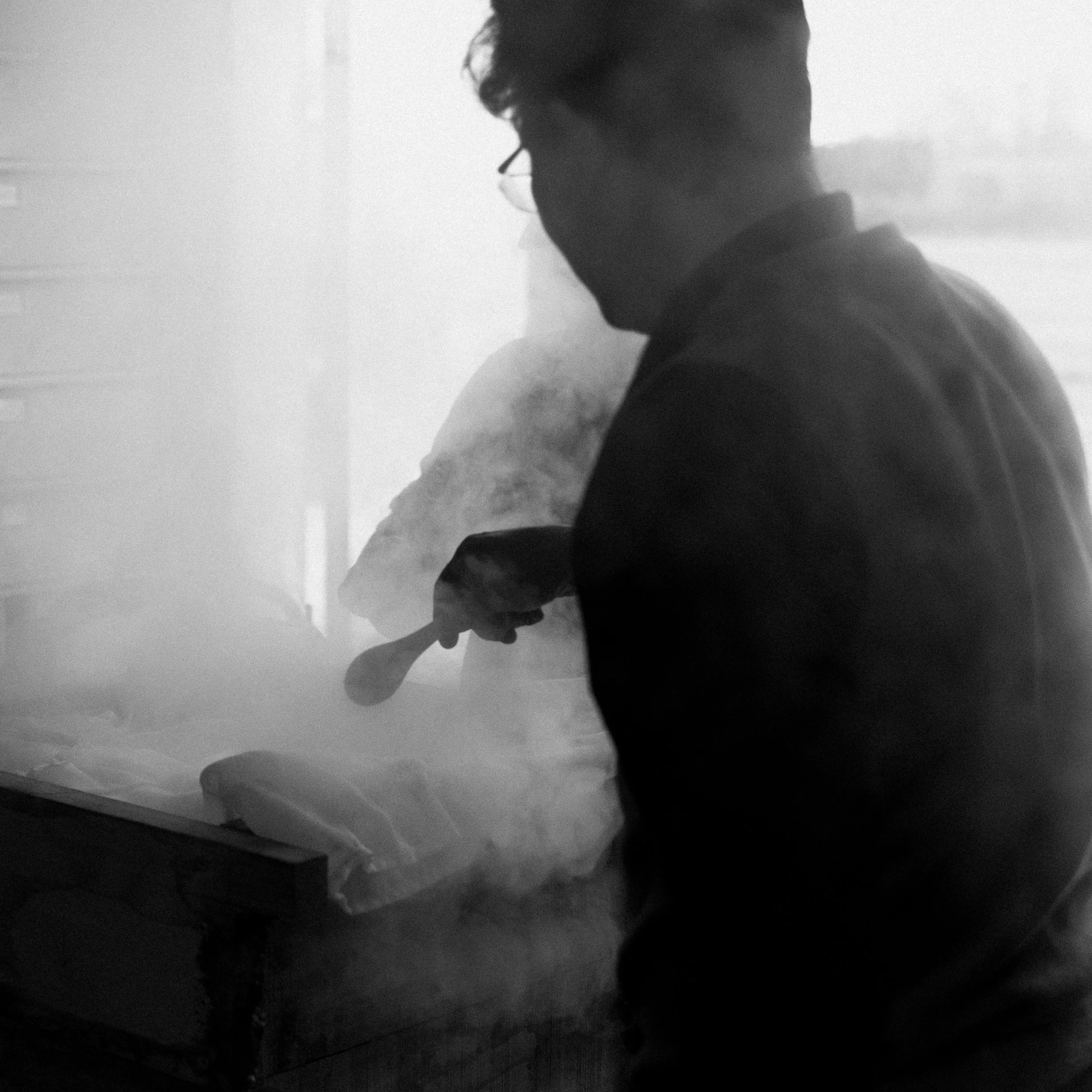 Une personne silhouette, portant des lunettes, en train de cuisiner dans une cuisine, avec de la vapeur visible, le tout en noir et blanc.
