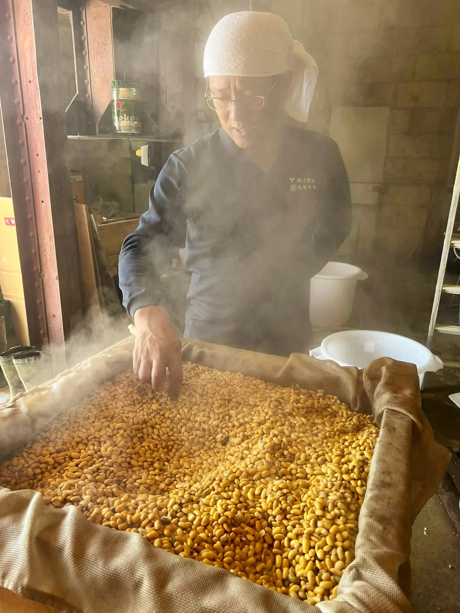Une personne portant un bandana blanc et des lunettes, qui travaille avec des graines jaunes dans un environnement de cuisine ou atelier, avec de la vapeur.