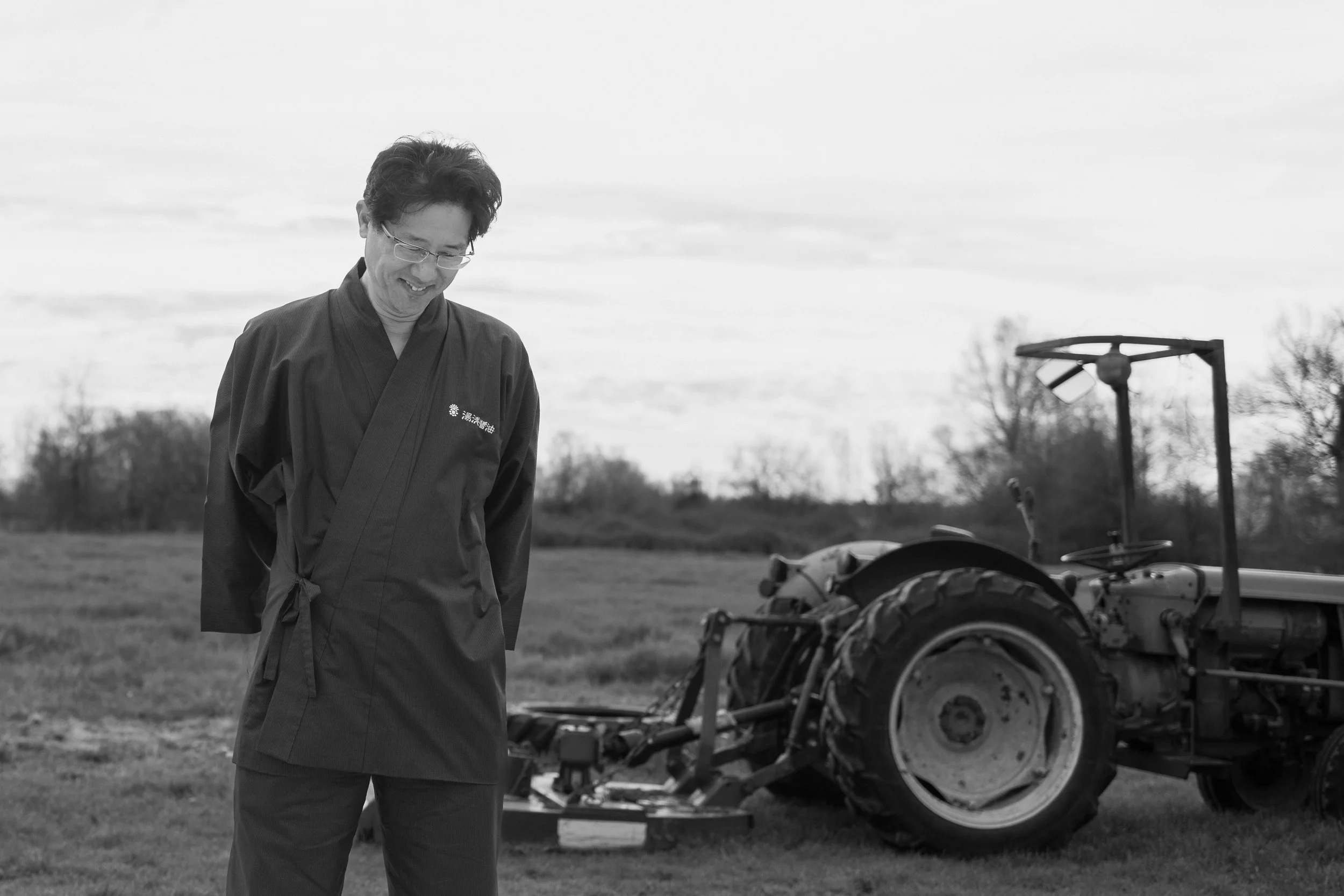 Un homme en vêtement traditionnel japonais, portant des lunettes, se tient dans un champ avec un tracteur en arrière-plan.
