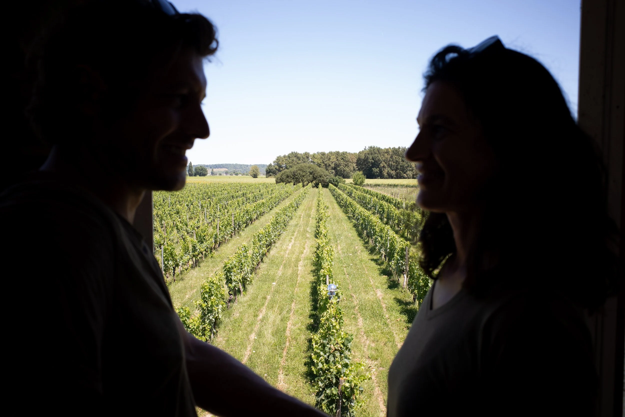 Deux personnes souriantes se tenant par la main devant une fenêtre qui donne sur un vignoble sous un ciel dégagé.