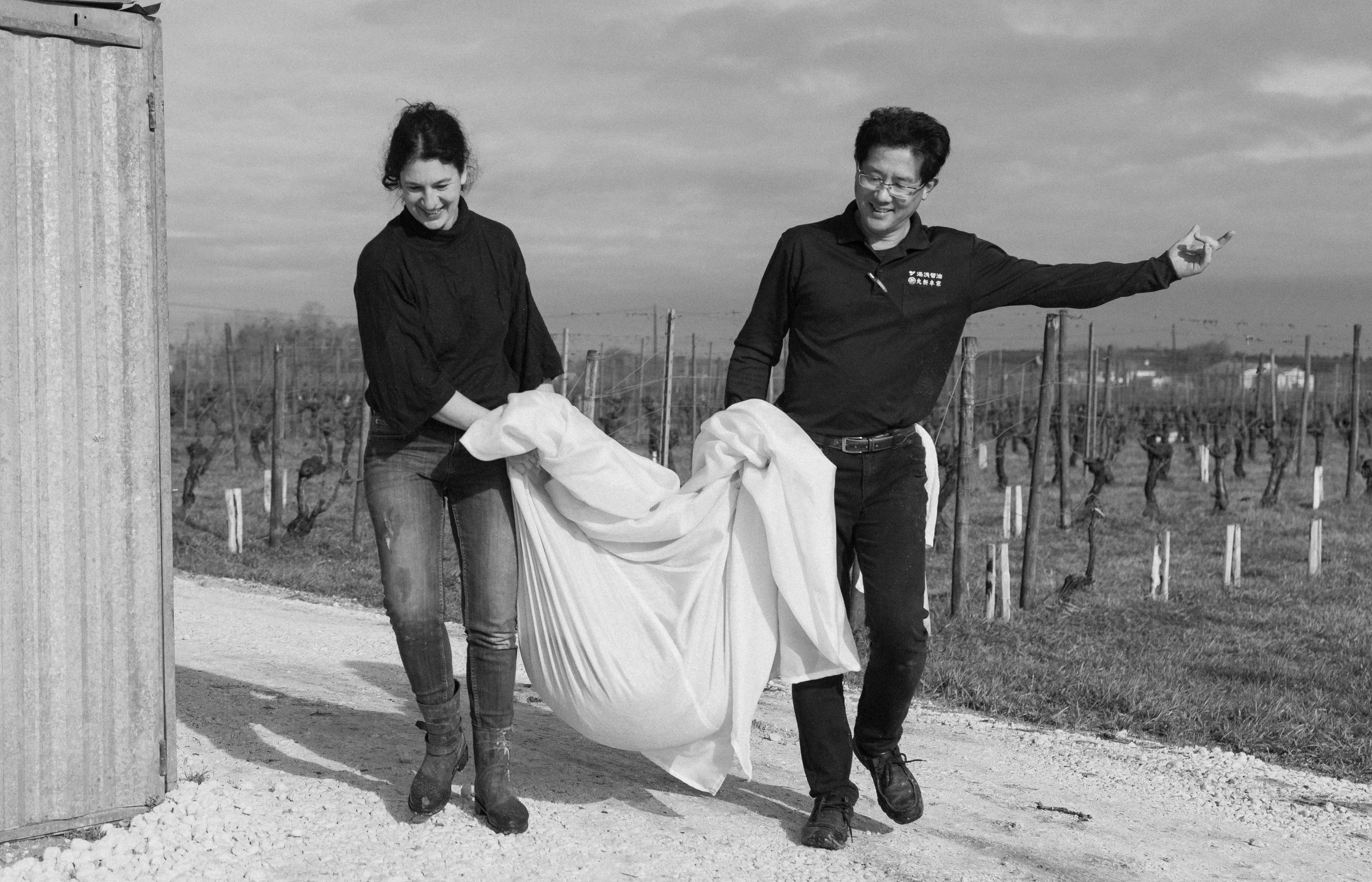 Deux personnes, un homme et une femme, qui sourient, transportent un sac blanc dans un vignoble.
