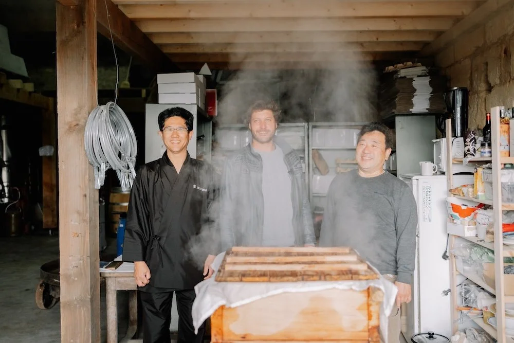Trois hommes souriants derrière une table en bois dans un atelier ou cuisine, avec de la vapeur qui monte, uns entres eux est Japonais, un autre est Européen, tous les trois posent pour la photo.