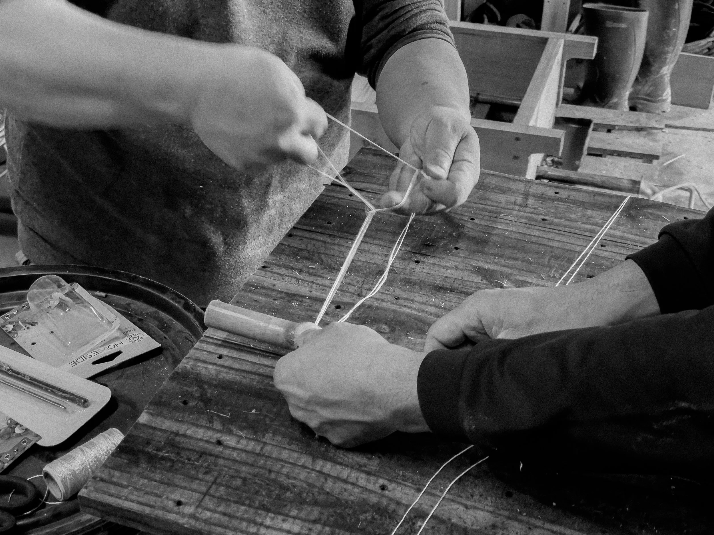Deux personnes tissent des filaments pour fabriquer un objet sur une table en bois avec des outils à proximité dans un atelier.