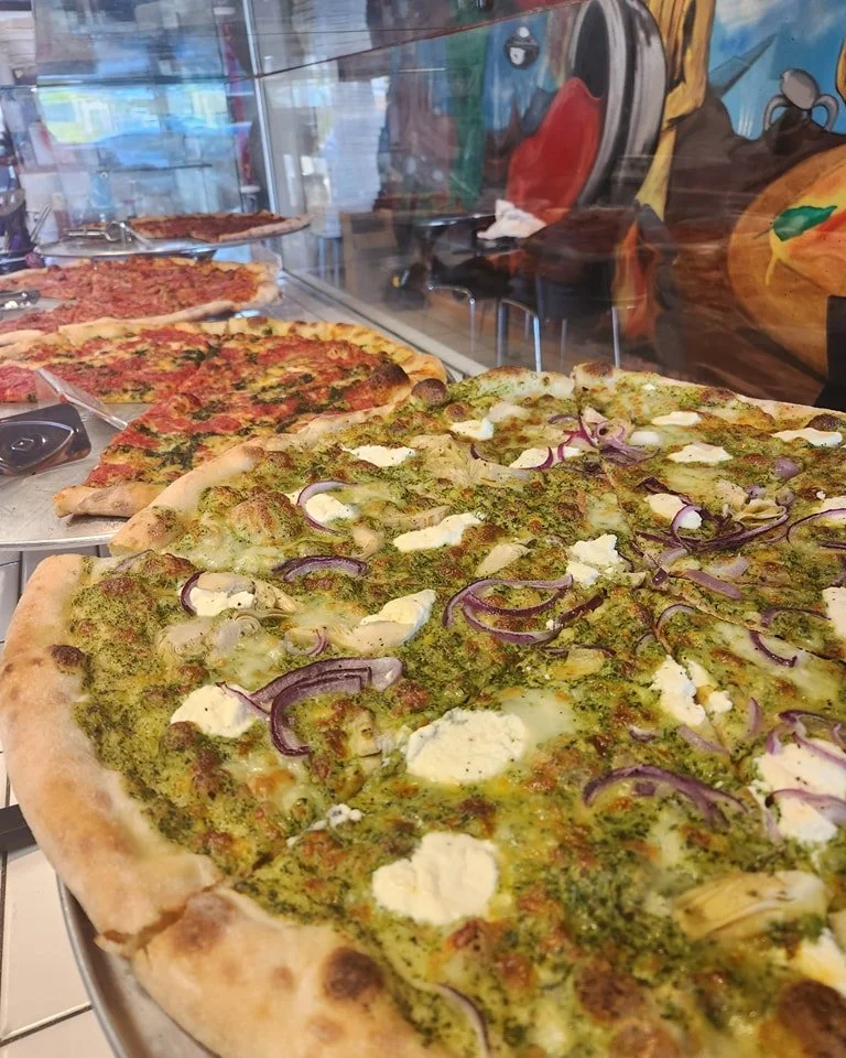 Close-up of a pesto pizza topped with cheese and red onions at a pizzeria display case.