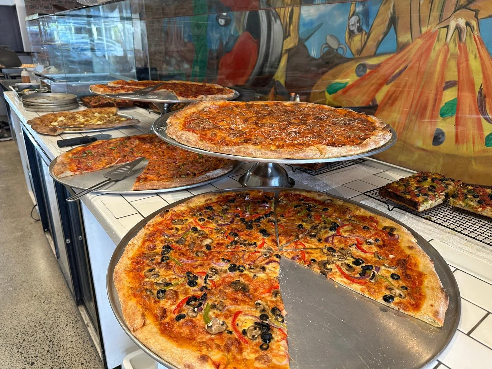 Assorted pizzas on display at a pizzeria, including a pepperoni, vegetable, and cheese pizza, with some slices removed.