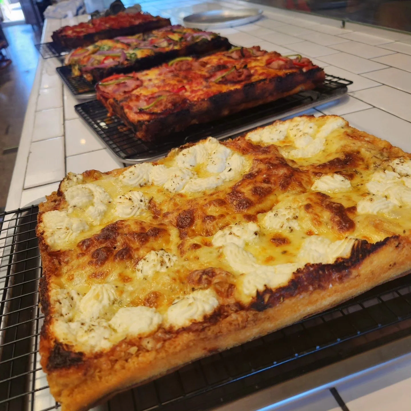 Three rectangular vegetarian pizzas cooling on wire racks, with the front pizza featuring cheese, dollops of ricotta, and herbs, and the two pizzas in the background topped with various vegetables.