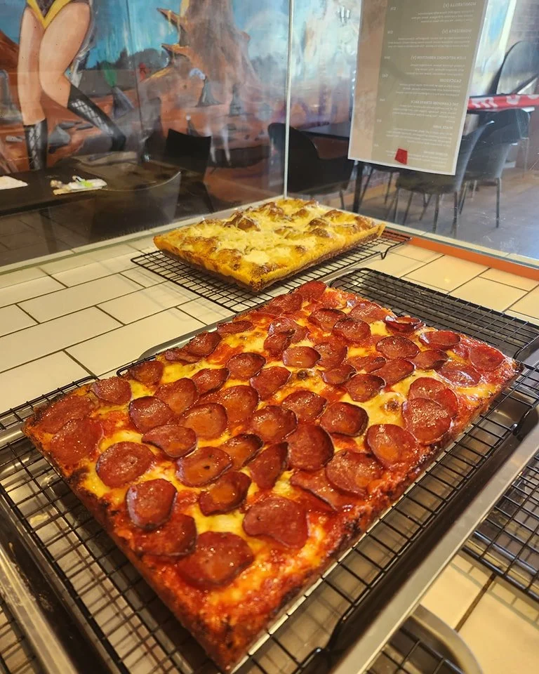 Two large rectangular pizzas on cooling racks inside a restaurant. The front pizza is topped with pepperoni and cheese, while the back pizza has a lighter topping.