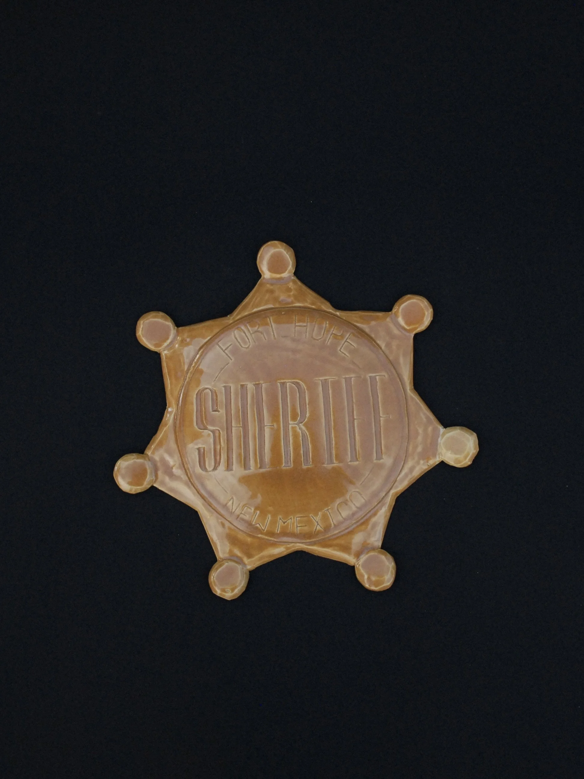 Image is of a ceramic sheriff's badge inspired by the Wild West, it features the inscription ‘Fort Hope, New Mexico Sheriff’ in raised lettering and is coloured all over in a warm yellow earthy glaze.