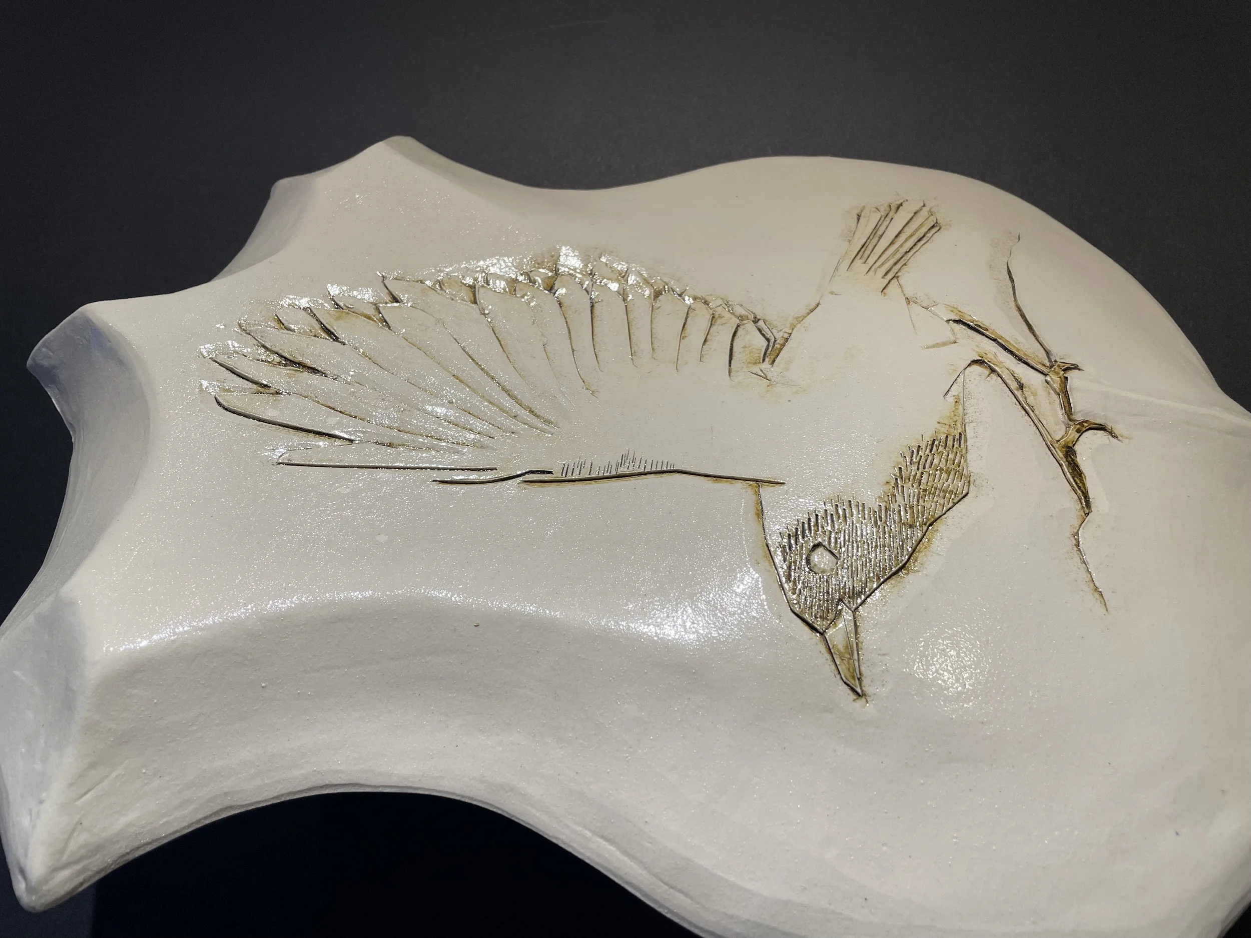 white ceramic shield-shaped wall piece carved with a bird on a branch, wing raised for flight, lines picked out in brown
