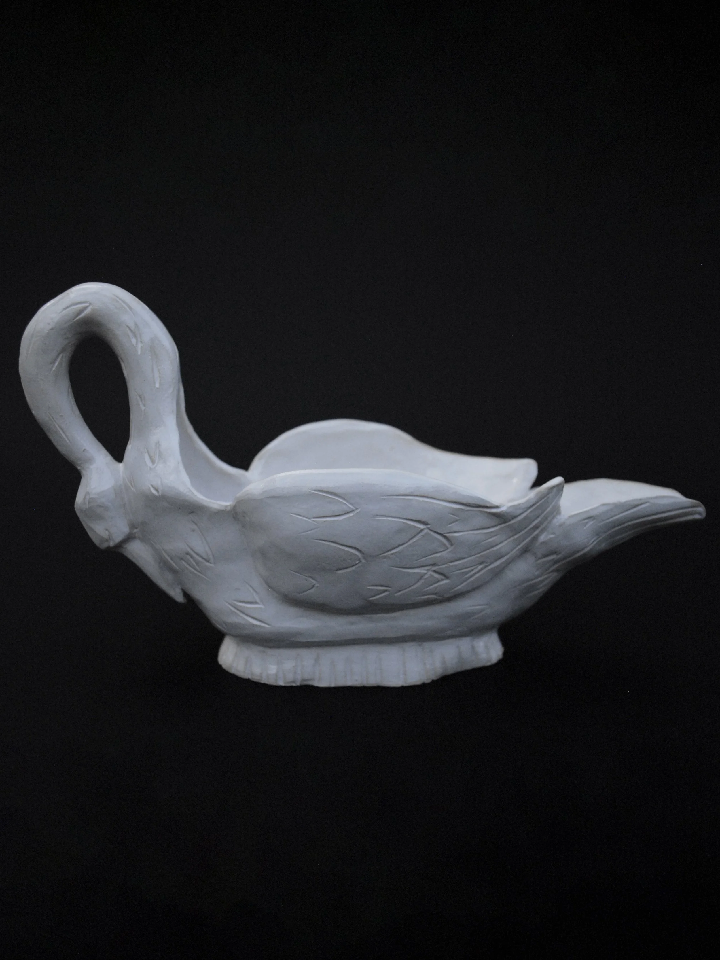 Image shows a ceramic hand-sculpted, swan shaped gravy boat, covered in white glaze and delicate details, it has carved feathers and a long neck which acts as the handle