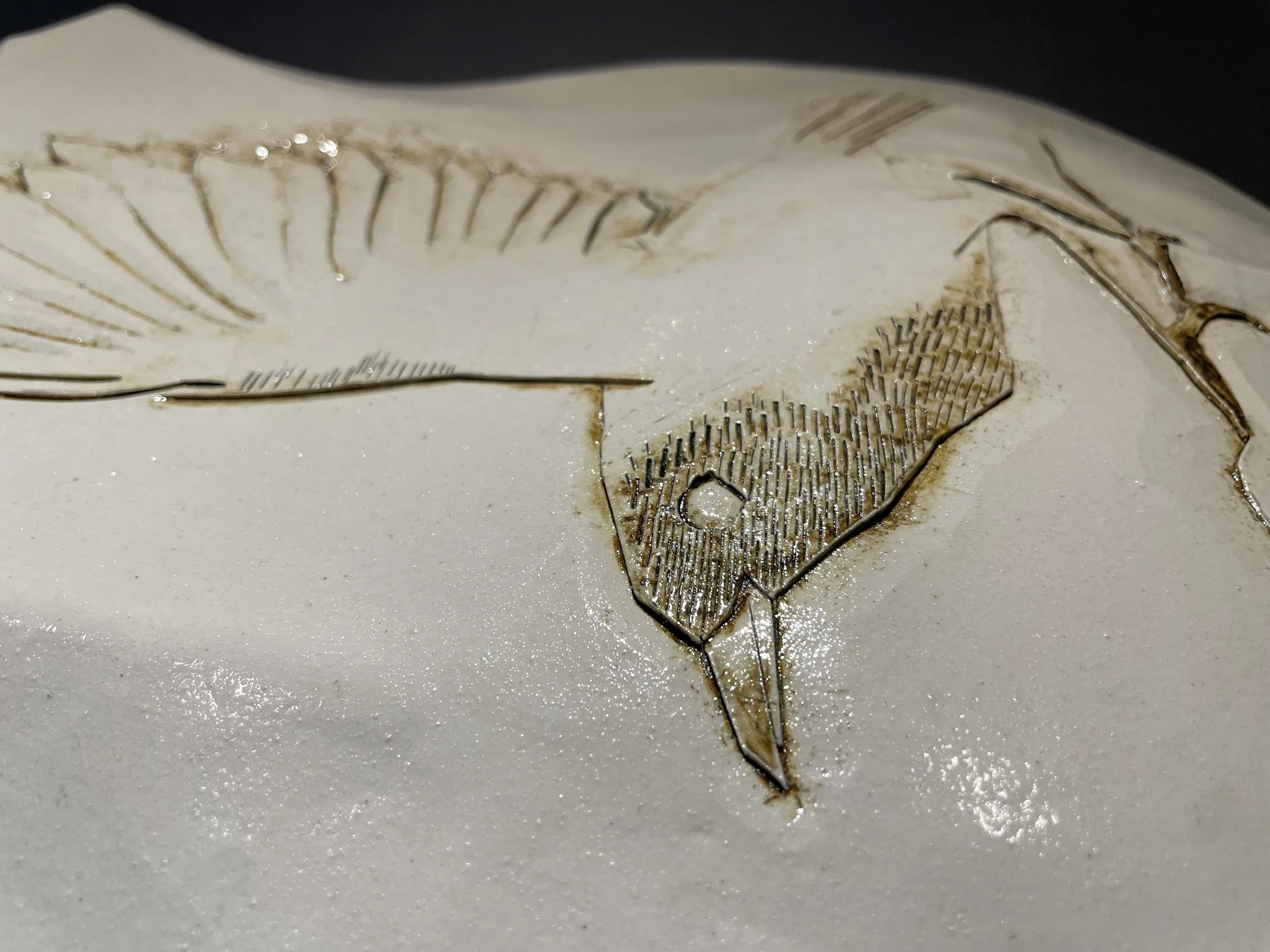 white ceramic shield-shaped wall piece carved with a bird on a branch, wing raised for flight, lines picked out in brown