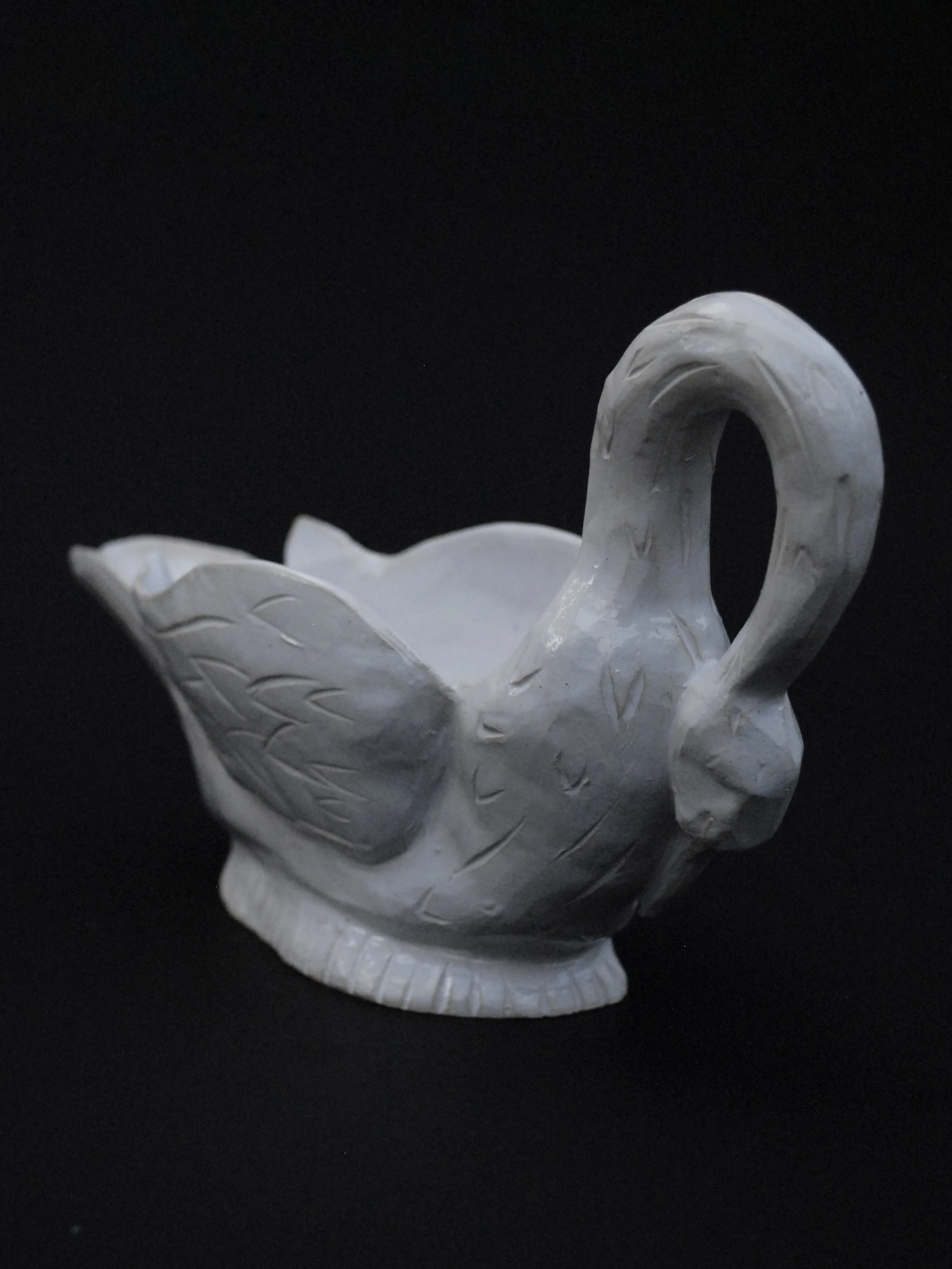 Image shows a ceramic hand-sculpted, swan shaped gravy boat, covered in white glaze and delicate details, it has carved feathers and a long neck which acts as the handle