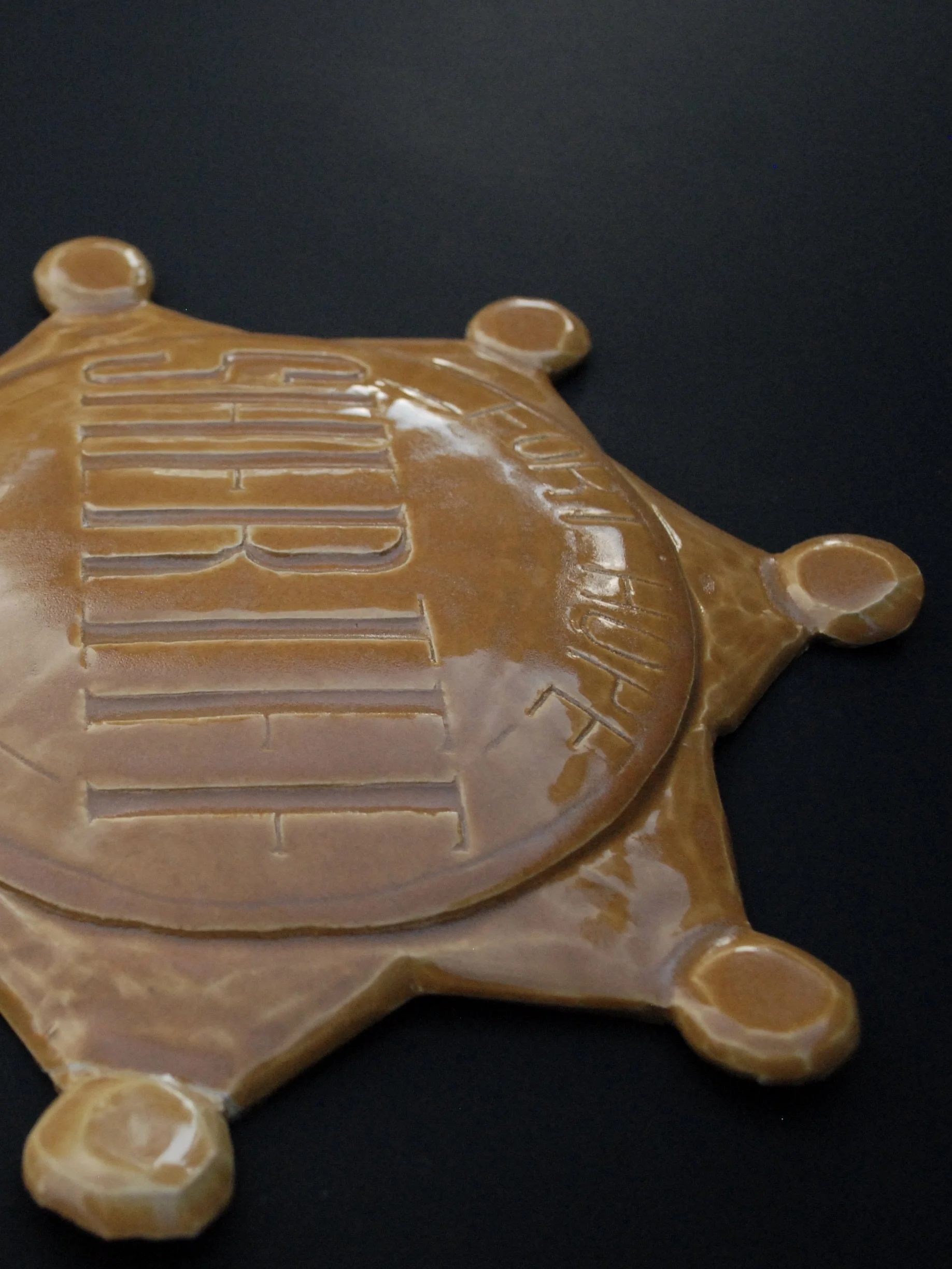 Image is of a ceramic sheriff's badge inspired by the Wild West, it features the inscription ‘Fort Hope, New Mexico Sheriff’ in raised lettering and is coloured all over in a warm yellow earthy glaze.