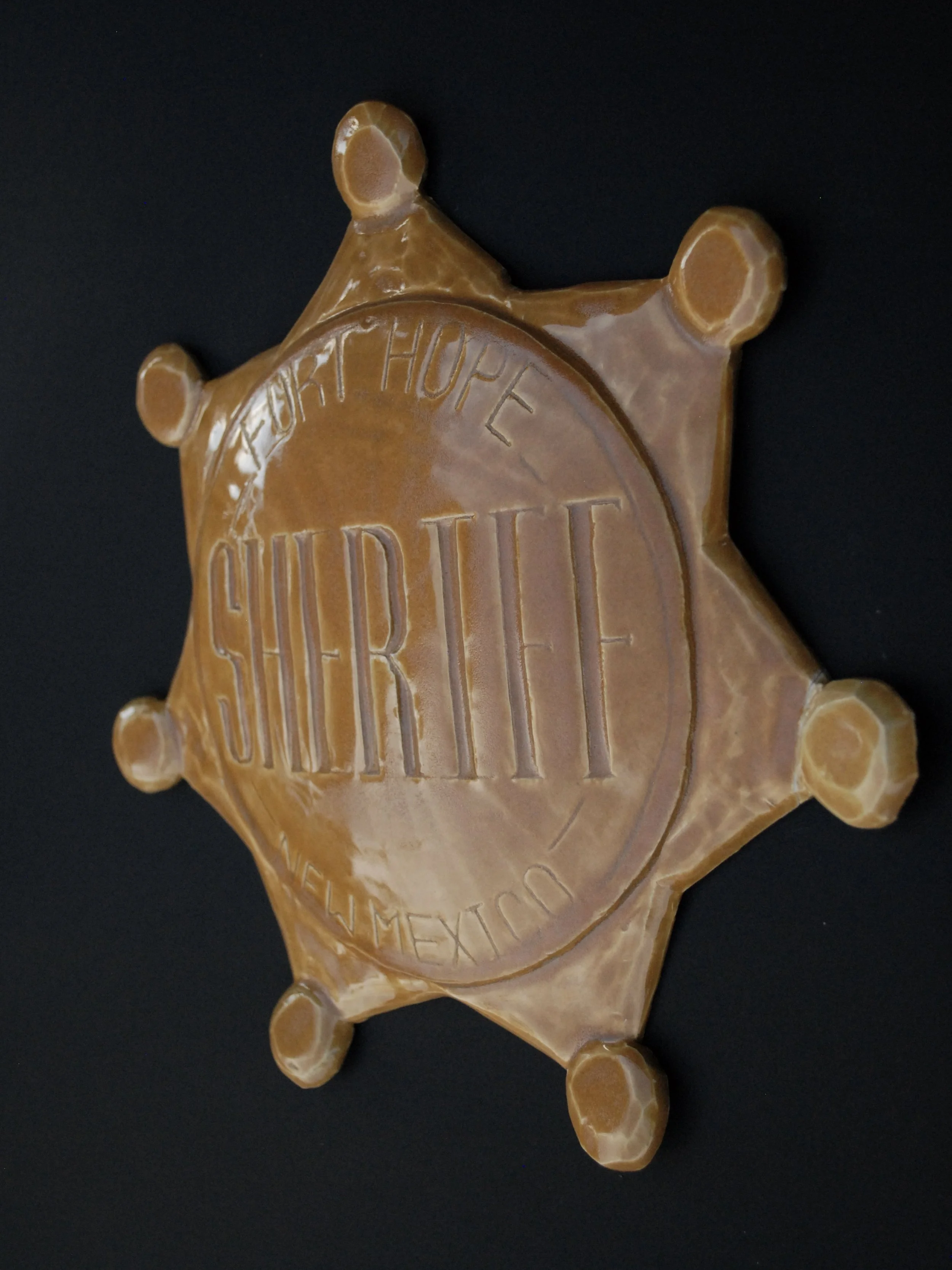 Image is of a ceramic sheriff's badge inspired by the Wild West, it features the inscription ‘Fort Hope, New Mexico Sheriff’ in raised lettering and is coloured all over in a warm yellow earthy glaze.
