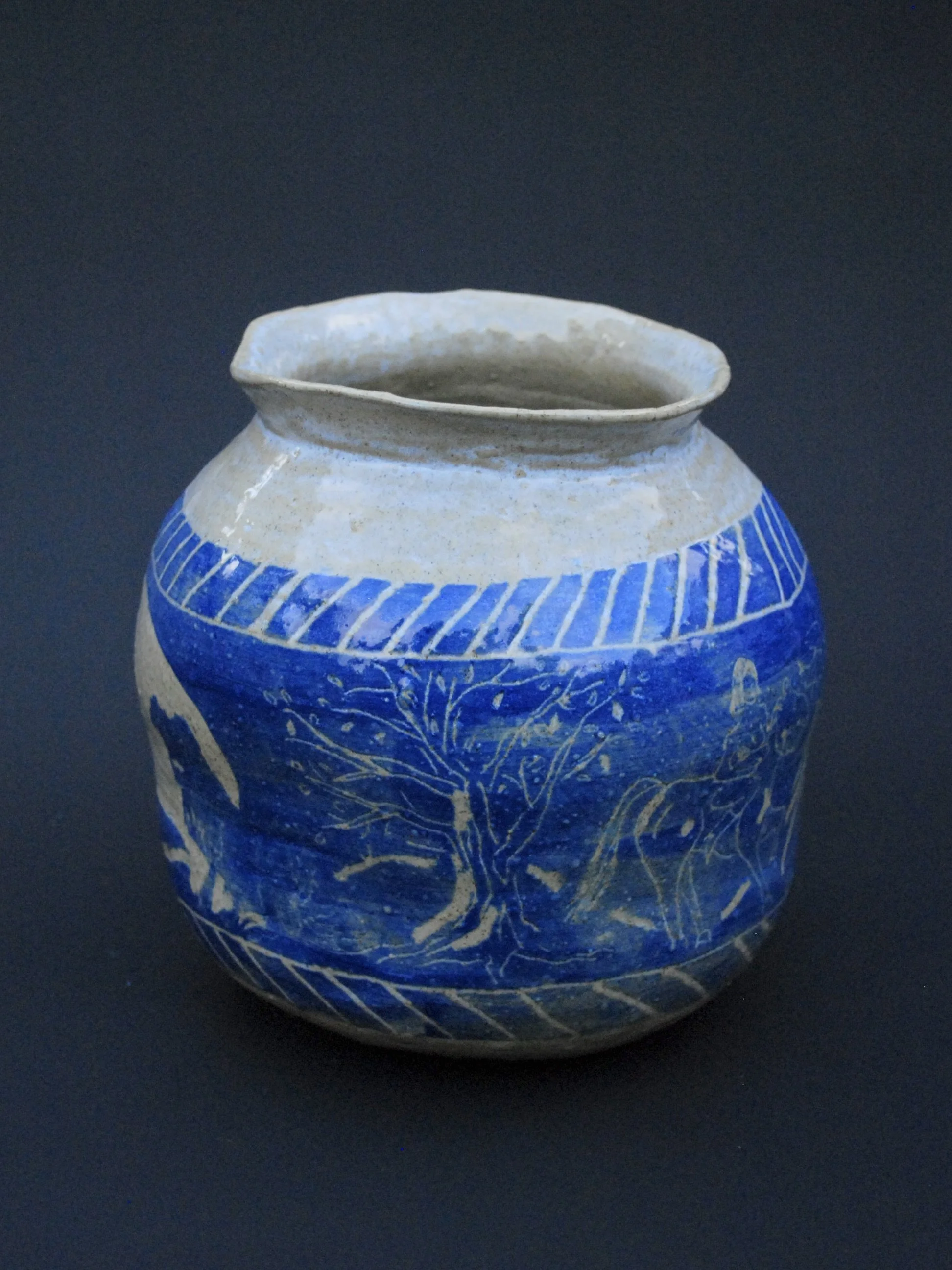A greek inspired stoneware pot with hand-etched blue designs against a warm, earthy tone. It has a unique and slightly irregular shape adding to the rustic appeal