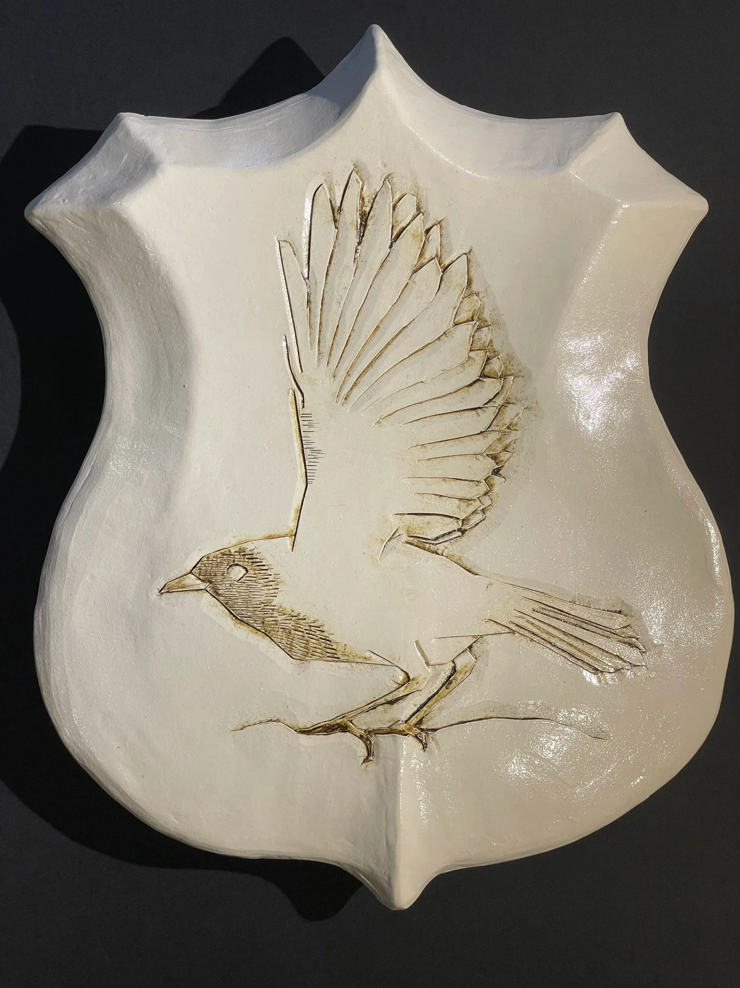white ceramic shield-shaped wall piece carved with a bird on a branch, wing raised for flight, lines picked out in brown