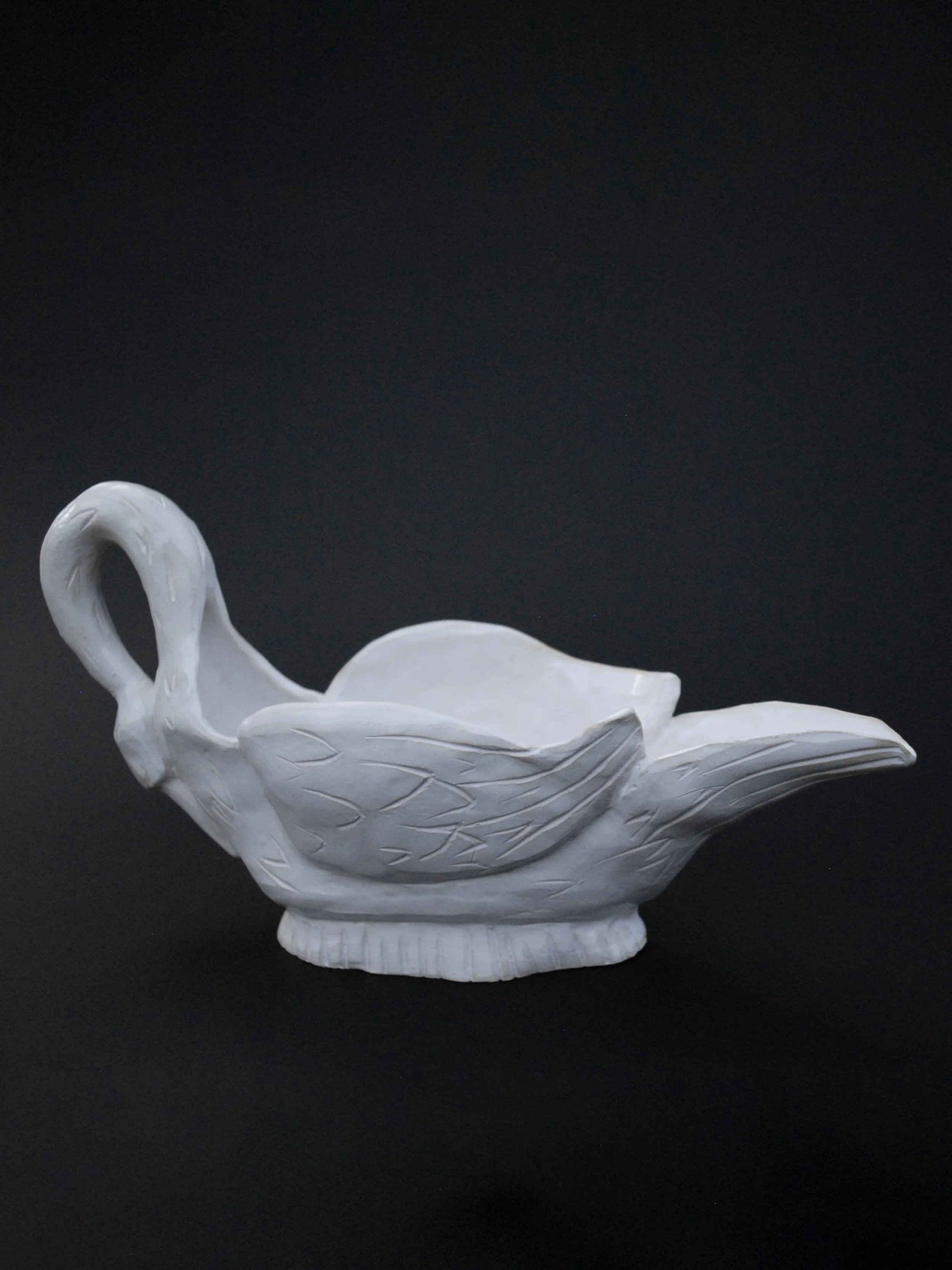 Image shows a ceramic hand-sculpted, swan shaped gravy boat, covered in white glaze and delicate details, it has carved feathers and a long neck which acts as the handle