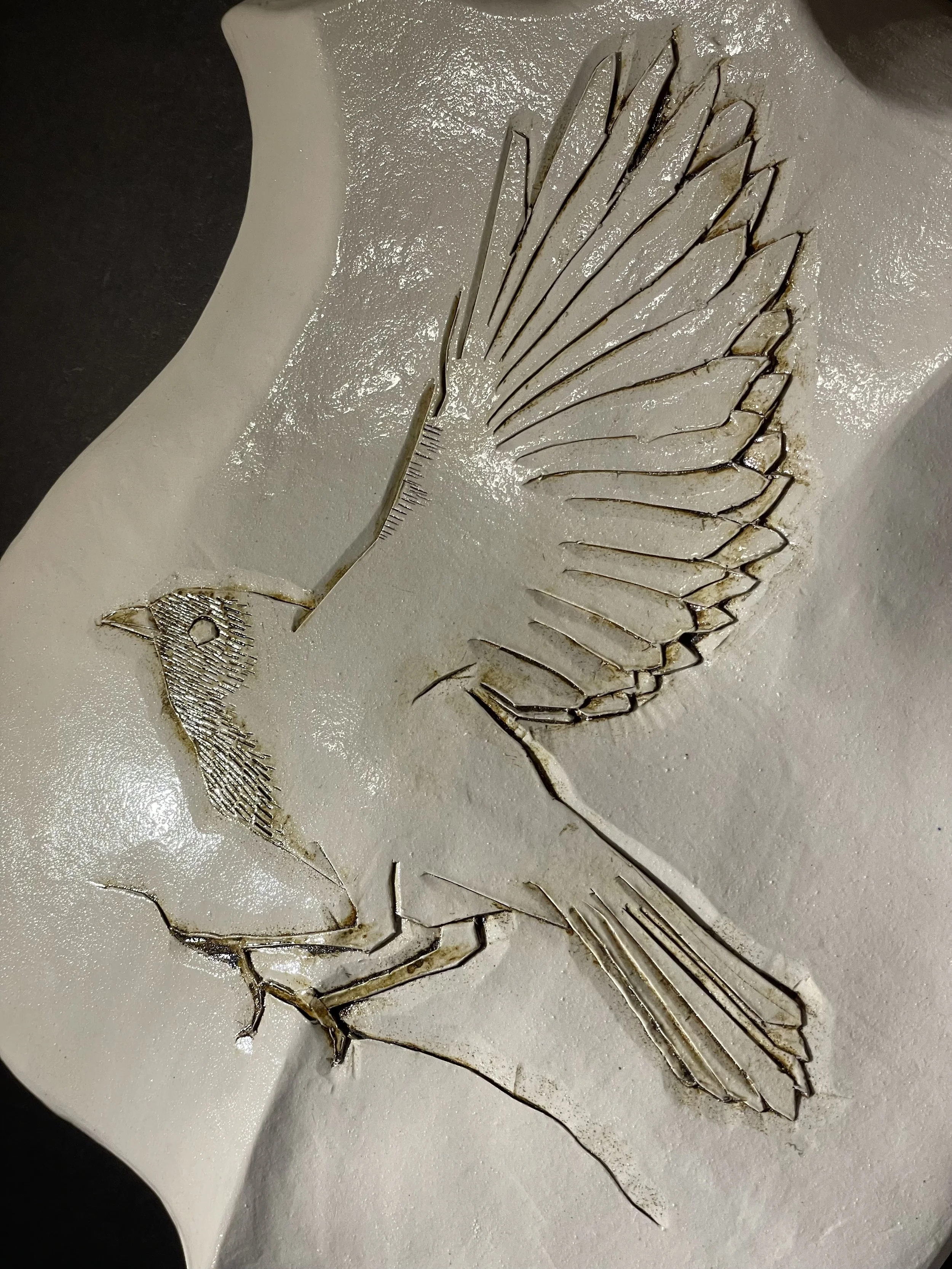 white ceramic shield-shaped wall piece carved with a bird on a branch, wing raised for flight, lines picked out in brown