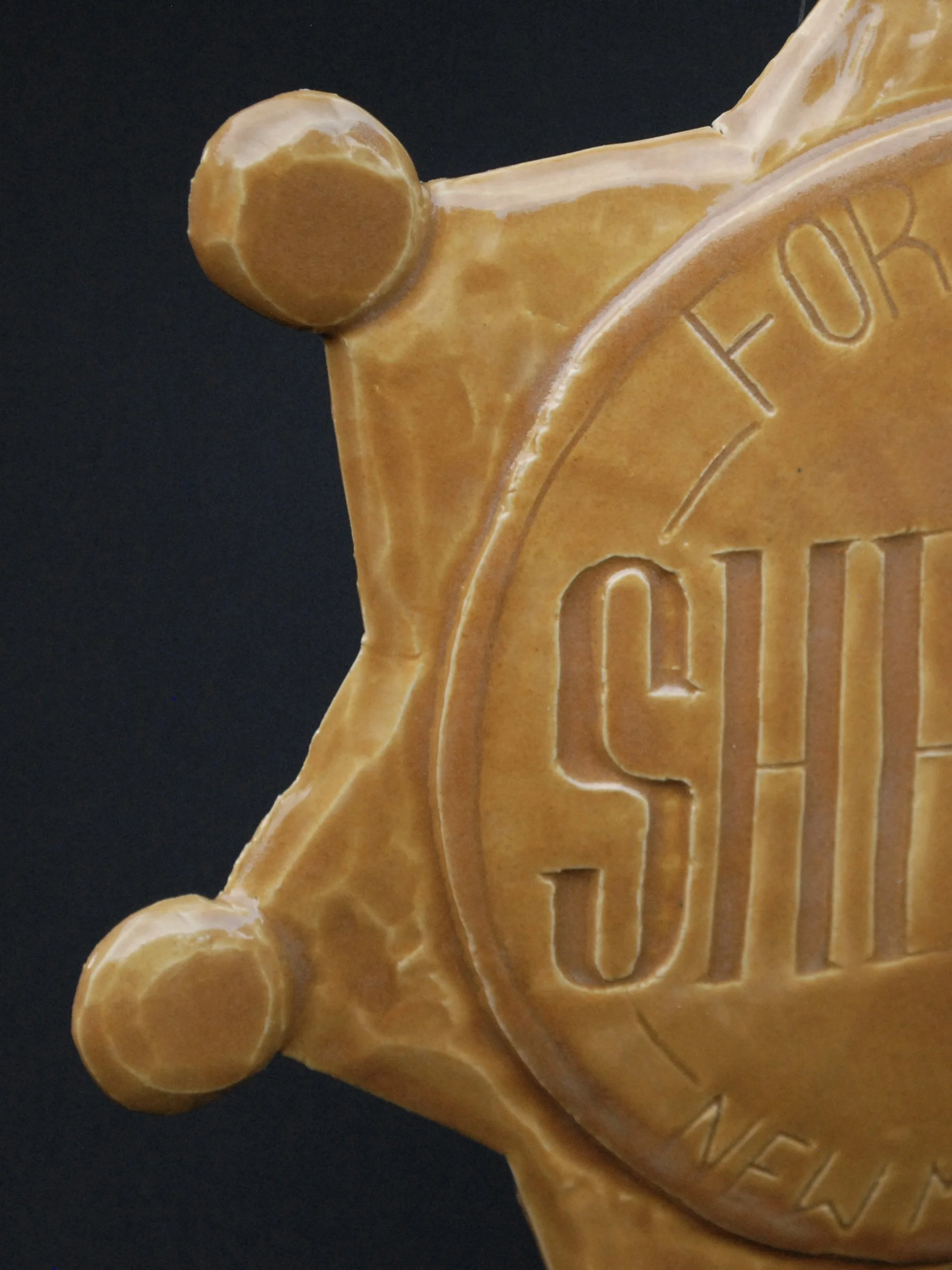 Image is of a ceramic sheriff's badge inspired by the Wild West, it features the inscription ‘Fort Hope, New Mexico Sheriff’ in raised lettering and is coloured all over in a warm yellow earthy glaze.