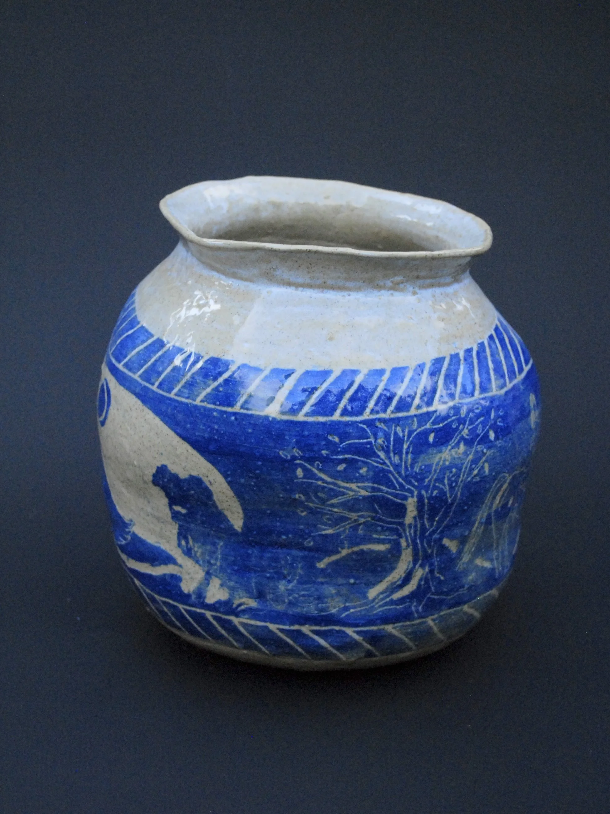A greek inspired stoneware pot with hand-etched blue designs against a warm, earthy tone. It has a unique and slightly irregular shape adding to the rustic appeal