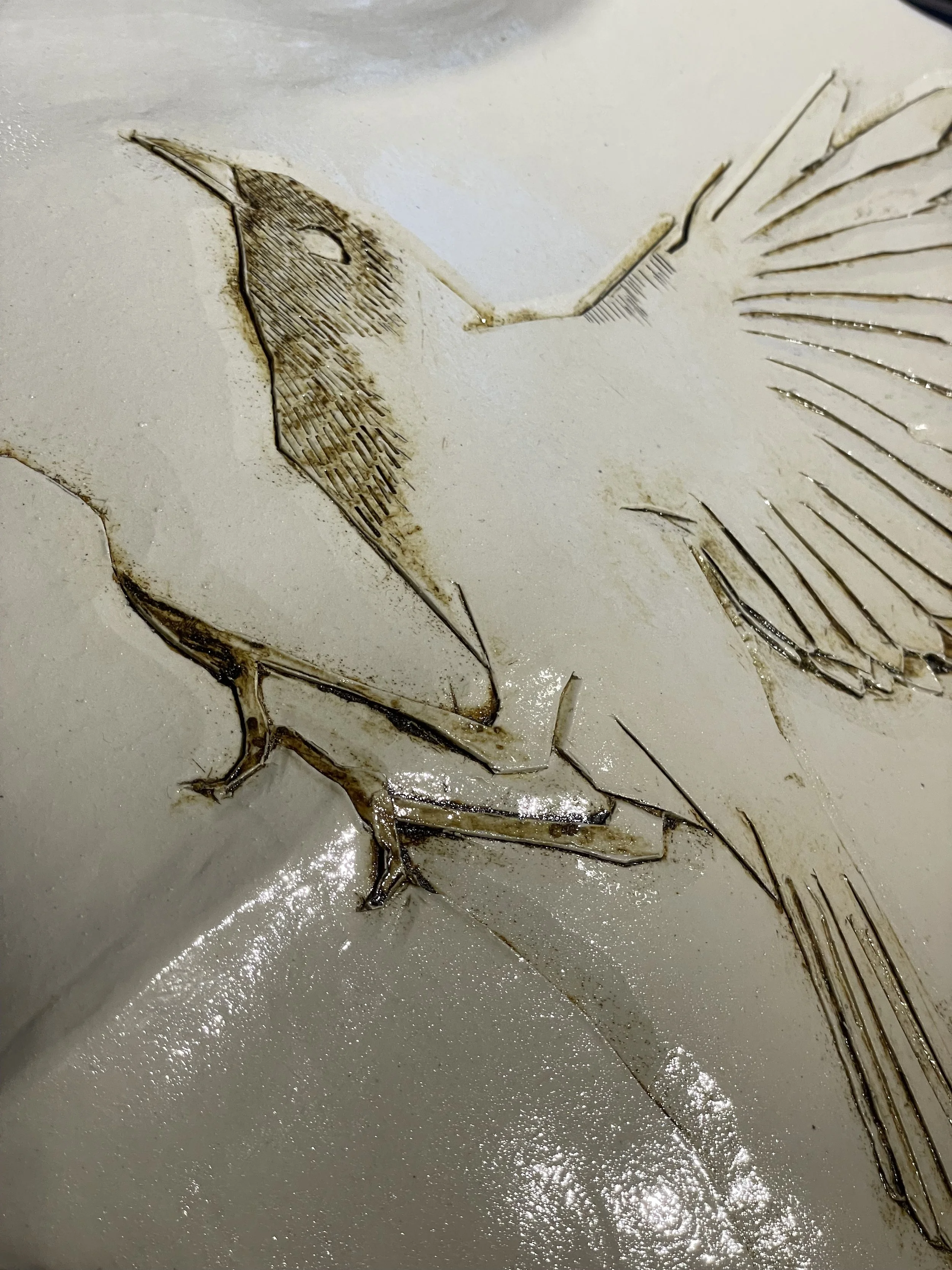 white ceramic shield-shaped wall piece carved with a bird on a branch, wing raised for flight, lines picked out in brown